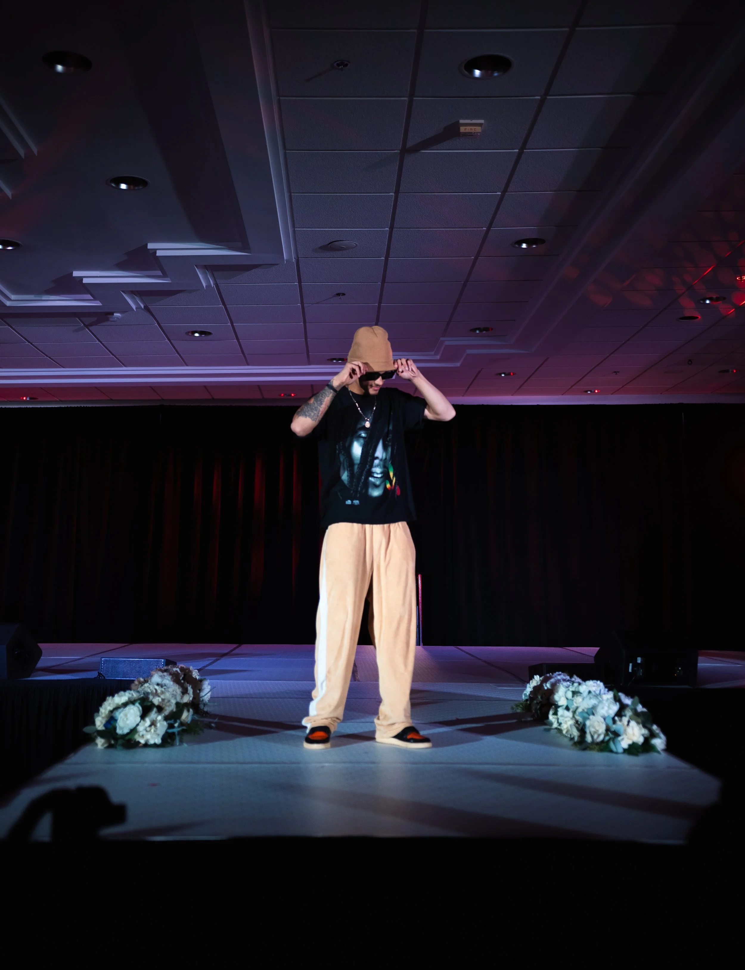 A man on a stage, wearing a tan hat, sunglasses, a black graphic t-shirt, tan pants, and sneakers, adjusting his hat, with flowers on the stage floor and a dark curtain behind him.