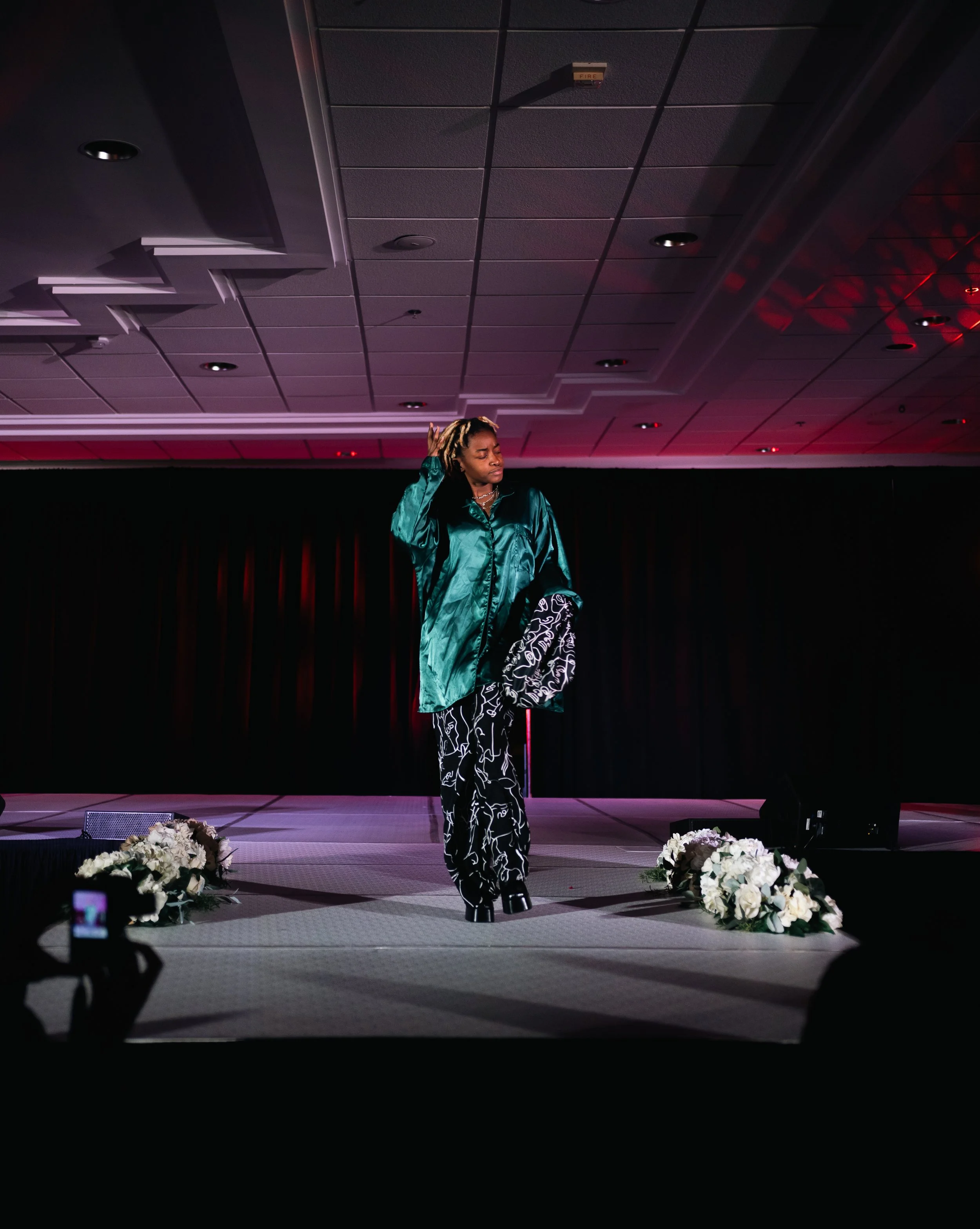 A woman standing on a stage with a black backdrop, floral arrangements on either side, wearing a shiny teal jacket and patterned pants, adjusting her hair while posing.