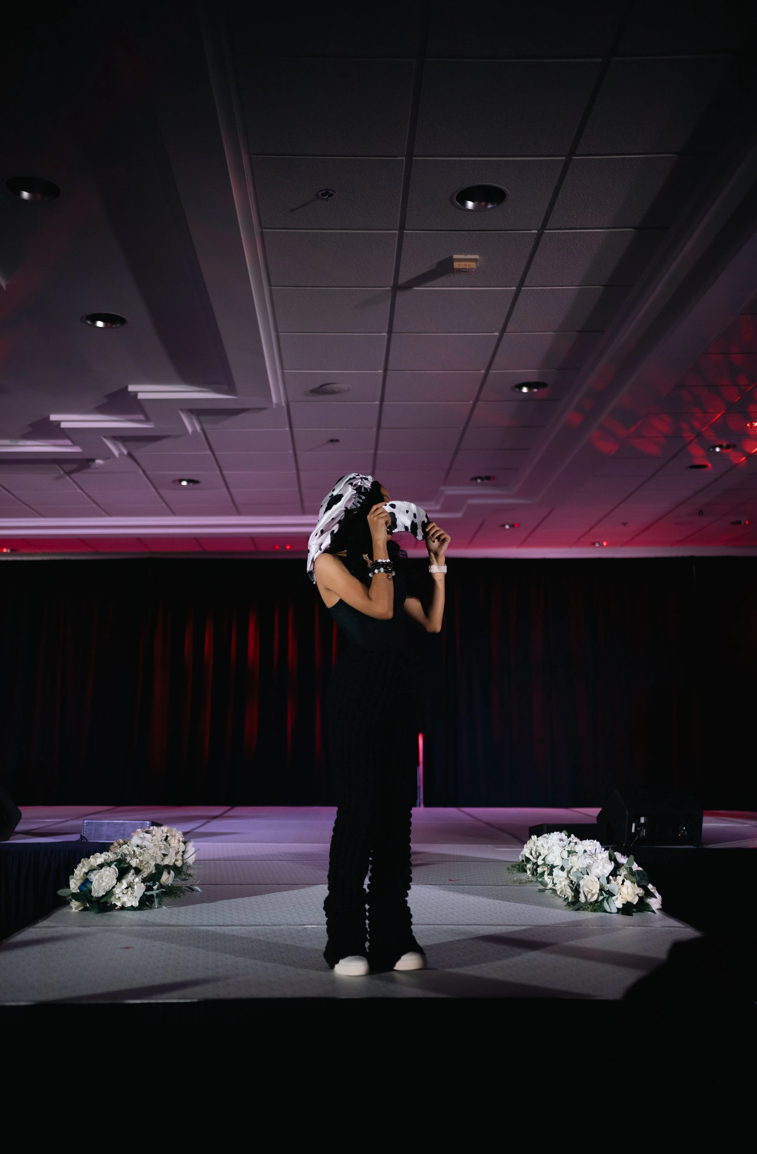 Person standing on stage, holding a cow-print hoodie over their head, surrounded by floral arrangements, in a dimly lit event space with black curtains.