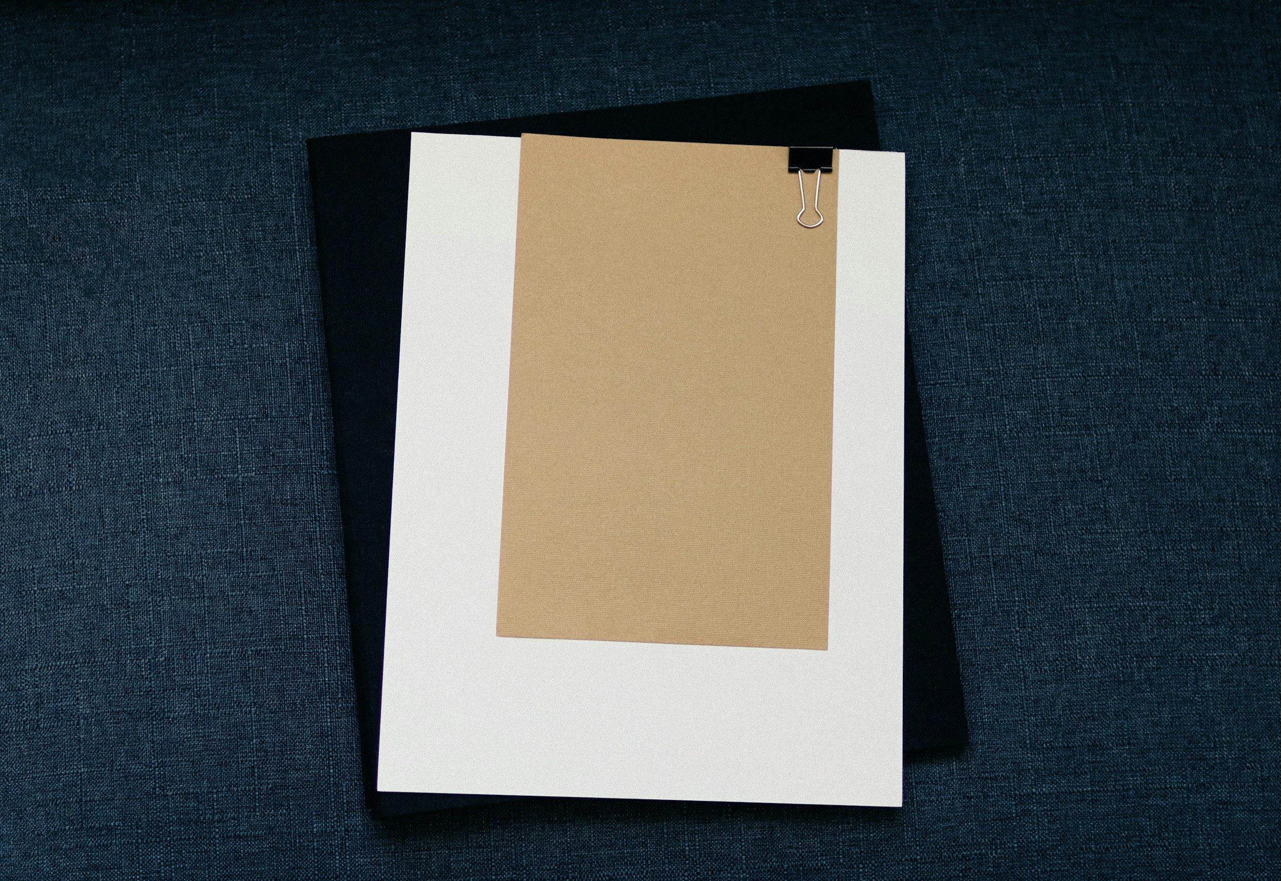 A white sheet of paper held with a black binder clip on a dark textured surface, with a beige sheet of paper attached on top of it.