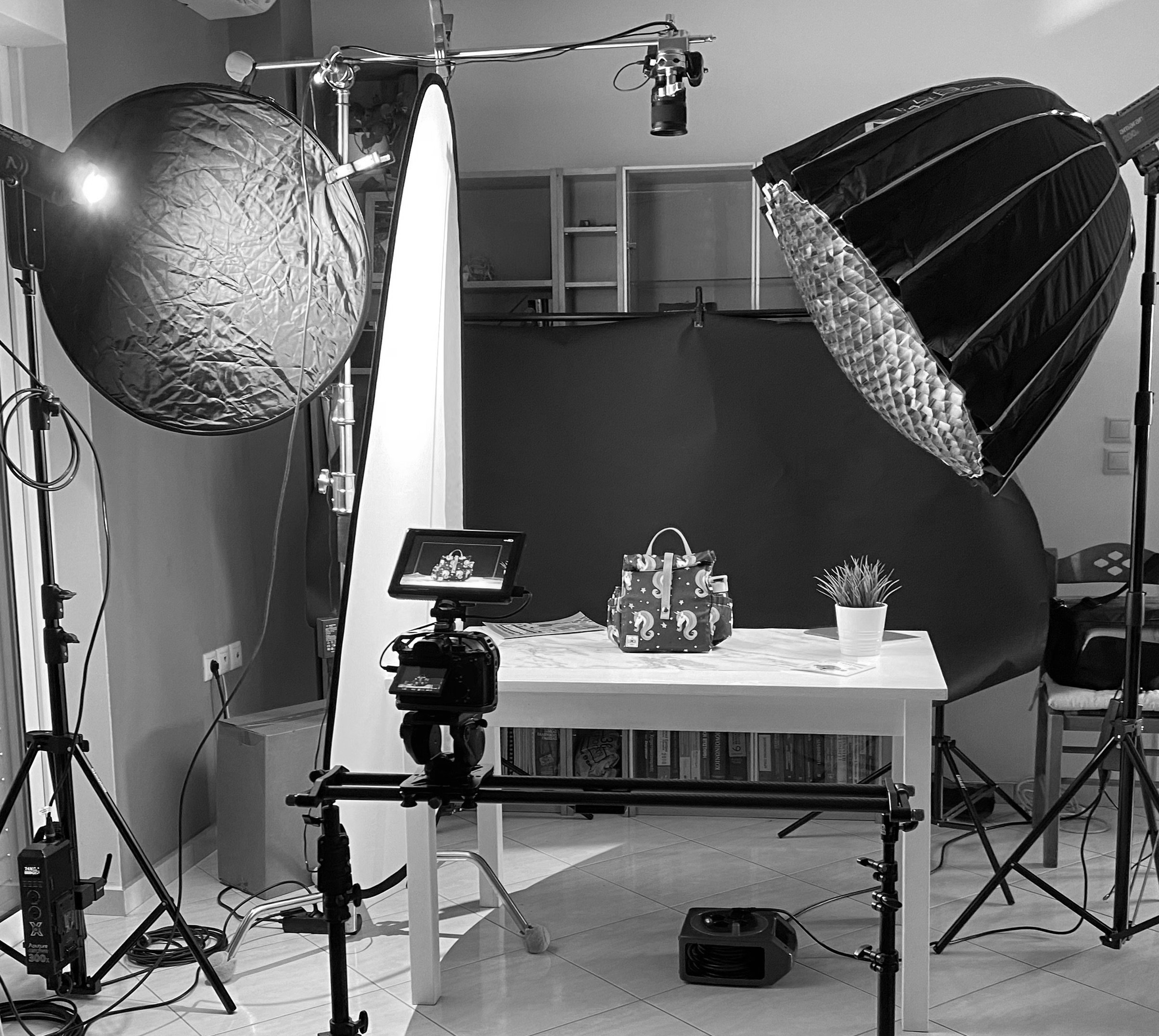 Photography studio setup with various lighting equipment, a computer monitor, a table with gifts, and bookshelves in the background.