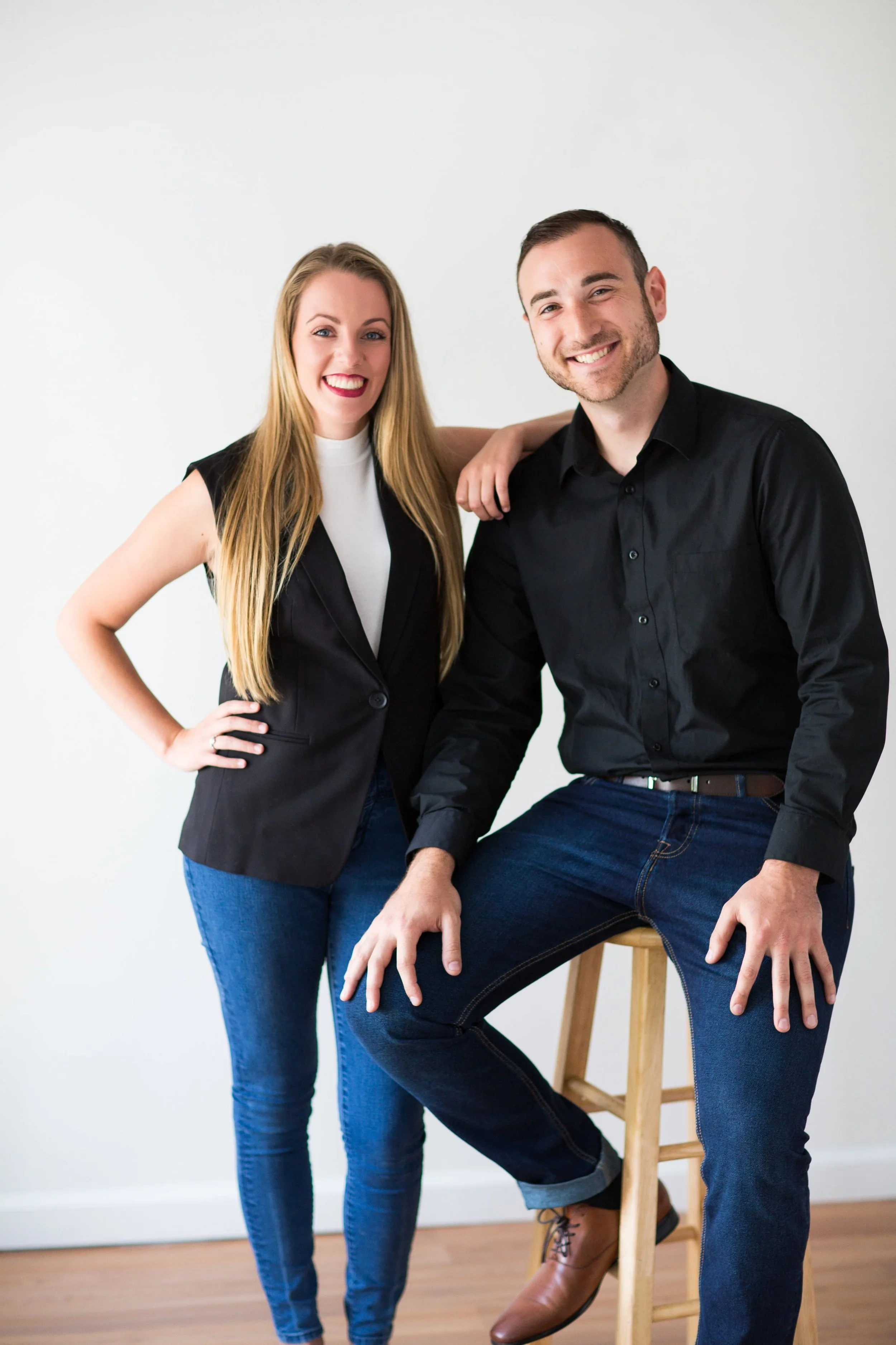 A woman with long blonde hair and a man with short dark hair smiling, with the woman standing and the man sitting on a wooden stool, both dressed in dark shirts and jeans, in front of a plain white background.