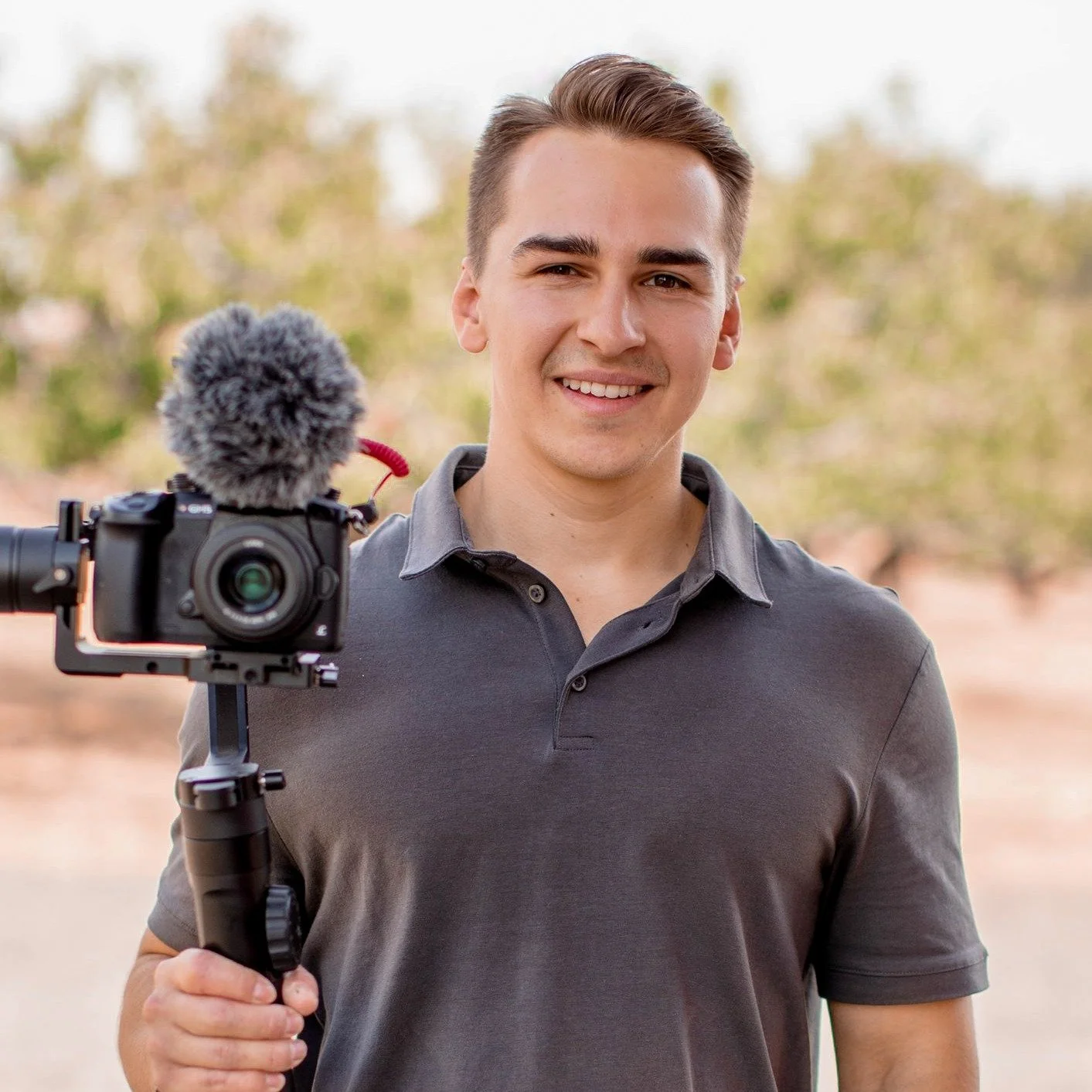 A young man smiling at the camera while holding a camera rig in an outdoor setting with trees in the background.