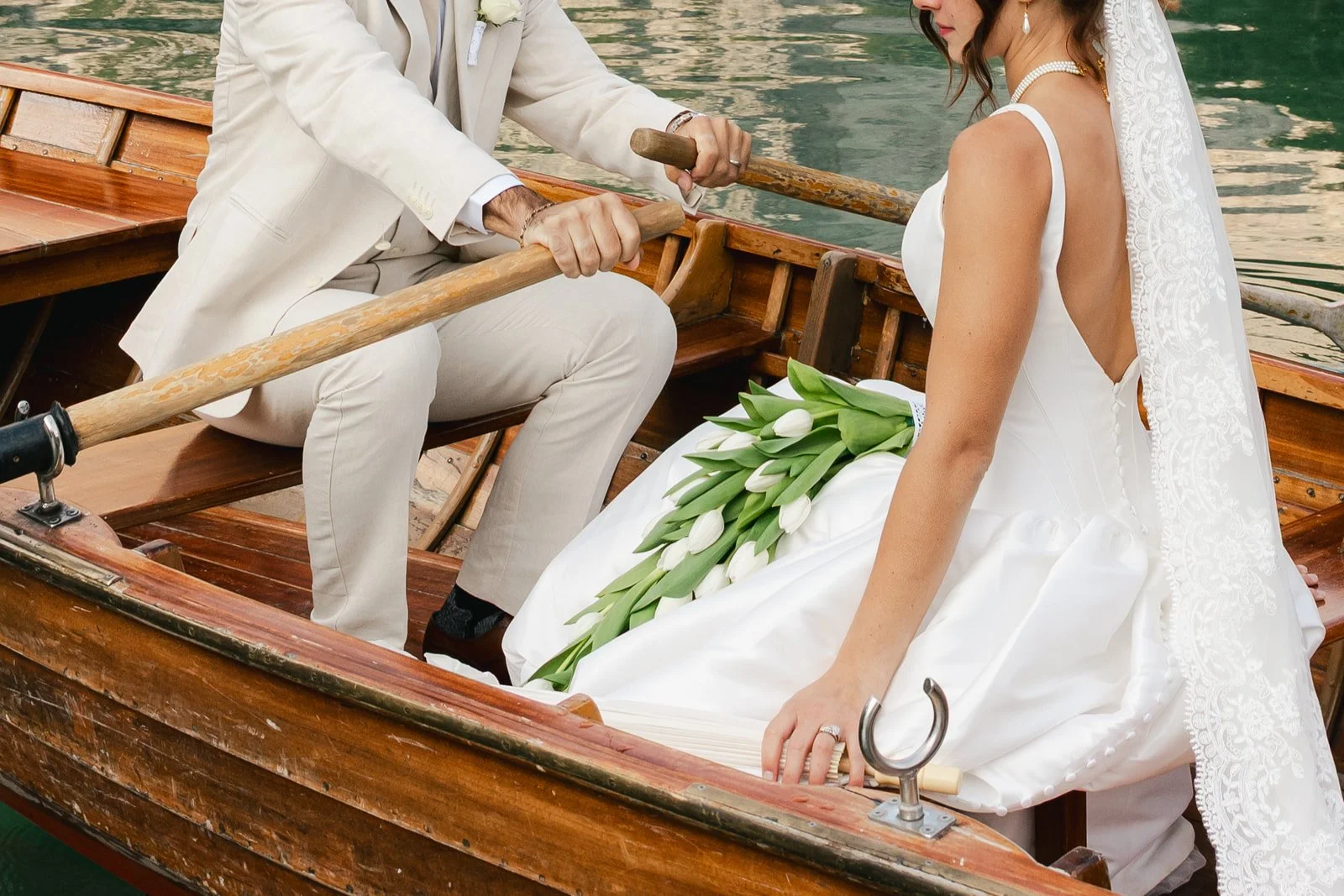 A bride in a white wedding dress sitting in a wooden boat, holding a bouquet of white tulips, with a man in a beige suit rowing the boat. Lago Di Braies in the Dolomites of Italy