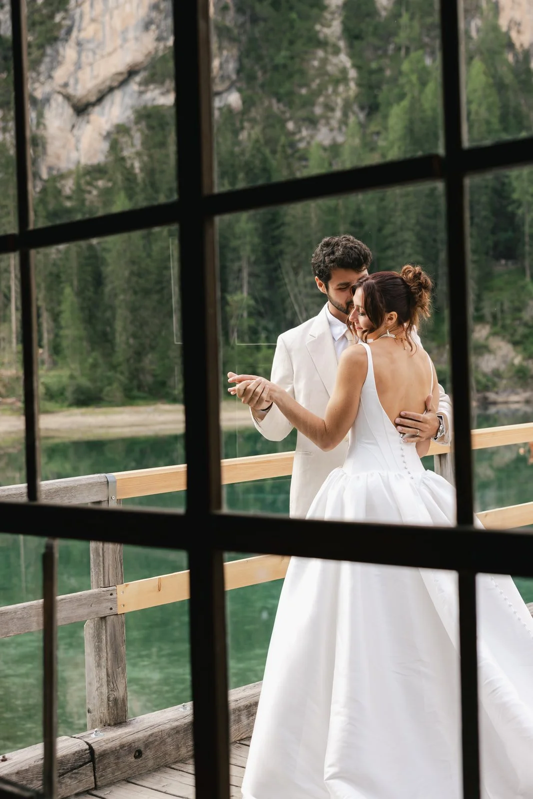 A couple in wedding attire dancing on a wooden deck near a lake, with forest and mountains in the background, seen through a window with black grid frames. Lago Di Braies in the Dolomites of Italy