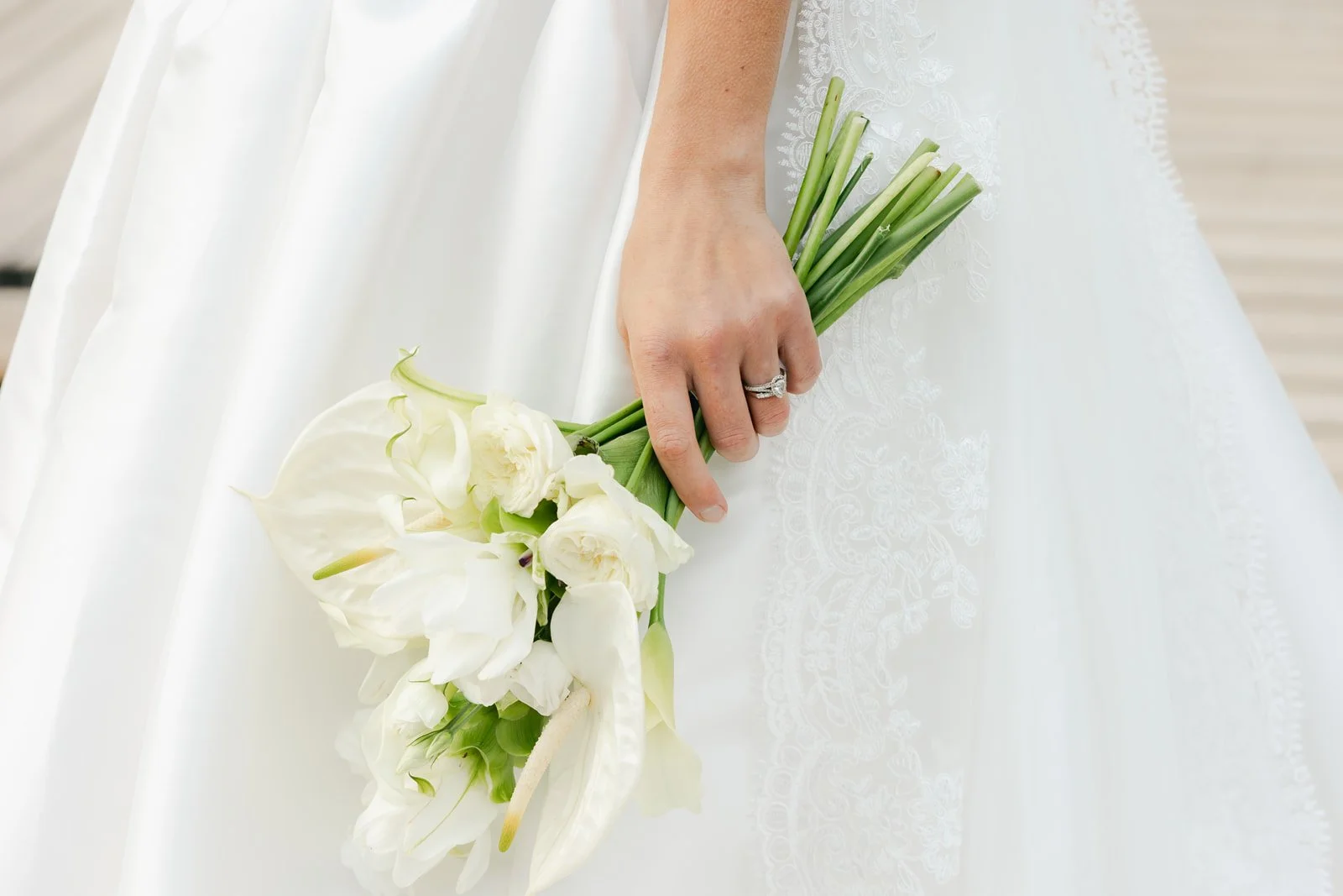A bride holding a bouquet of white flowers, including lilies and roses, with a wedding ring on her finger, against a white lace dress. Lago Di Braies in the Dolomites of Italy