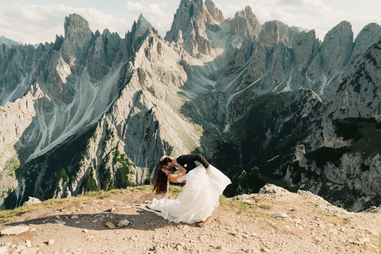 A couple in wedding attire sharing a romantic kiss on a mountain outcrop with towering rocky mountains in the background.