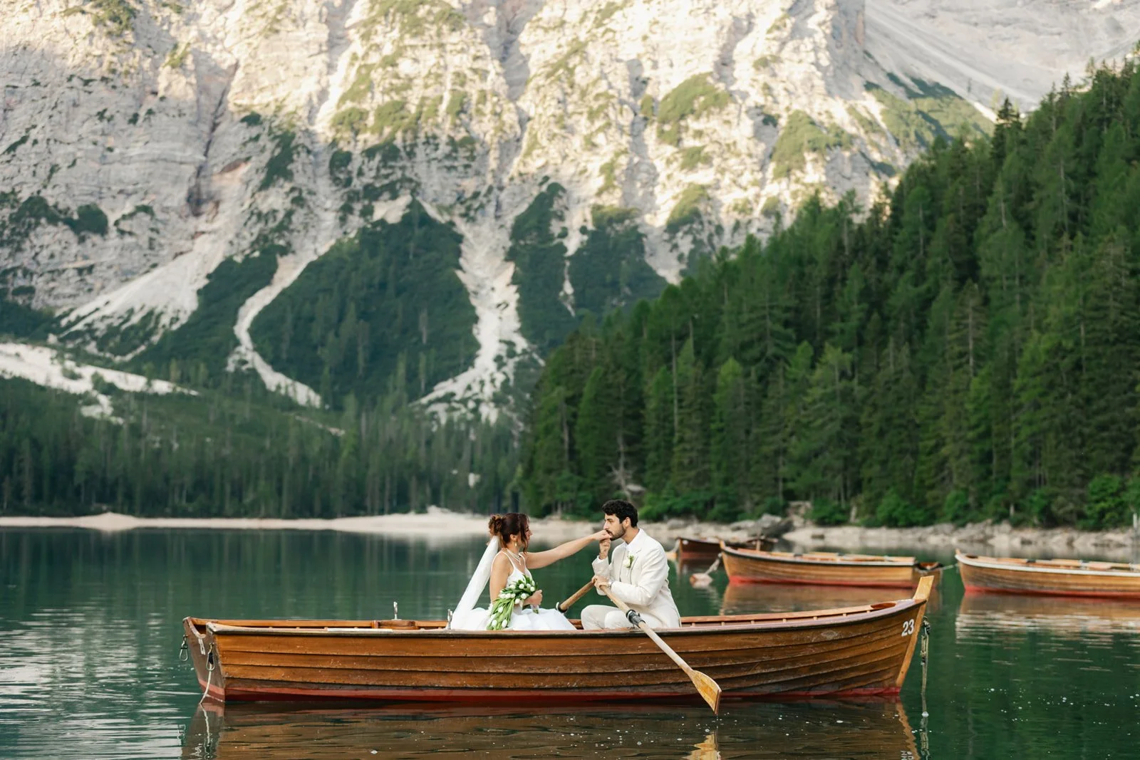 A bride and groom sitting in a wooden boat on a calm lake surrounded by mountains and dense green trees, with several other boats in the background. Lago Di Braies in the Dolomites of Italy