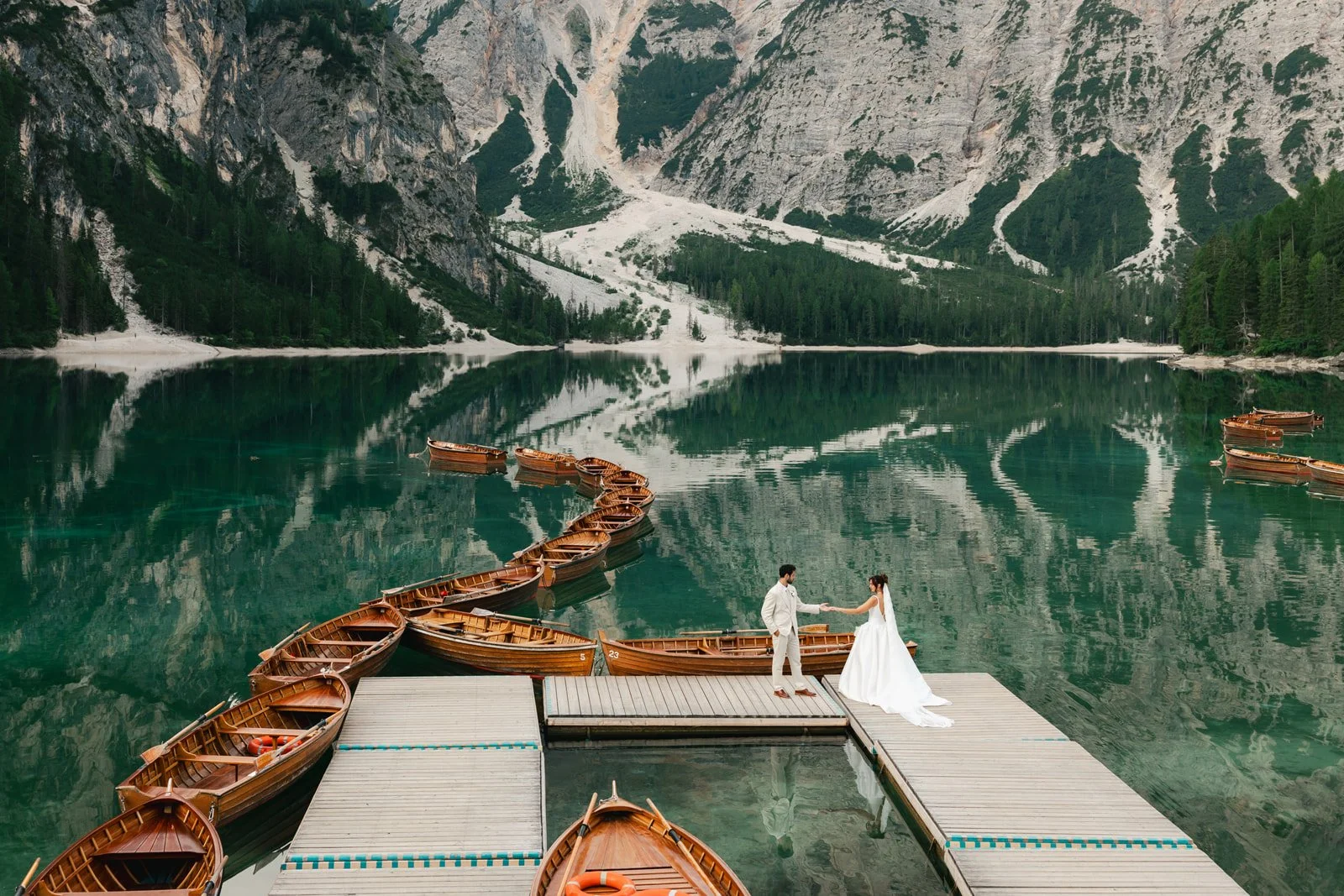 A bride and groom shake hands on a dock surrounded by wooden boats, with a mountain lake and forest in the background. Lago Di Braies in the Dolomites of Italy
