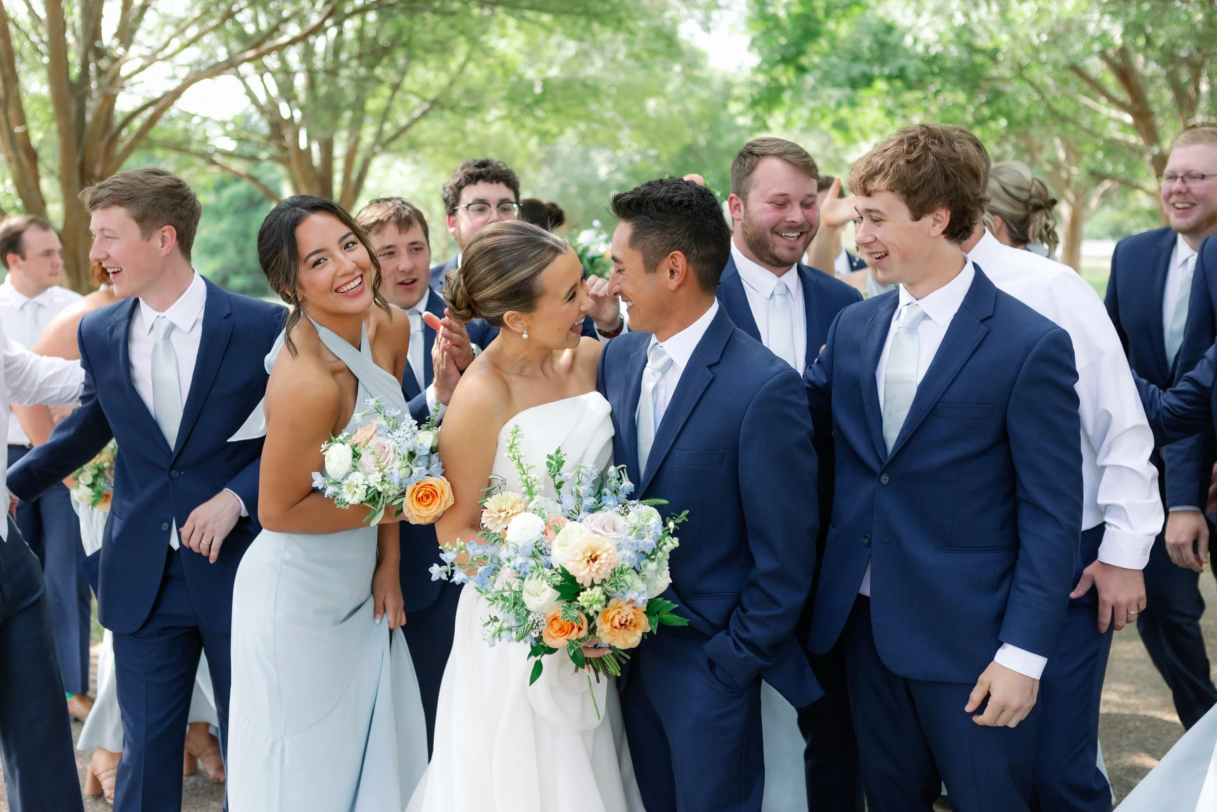 The bridal party showcases cohesive floral design with pastel blue accents, textured white blooms, and warm peach details. This group portrait highlights the romantic summer palette and refined floral styling created for Callie & Austin’s Missouri we