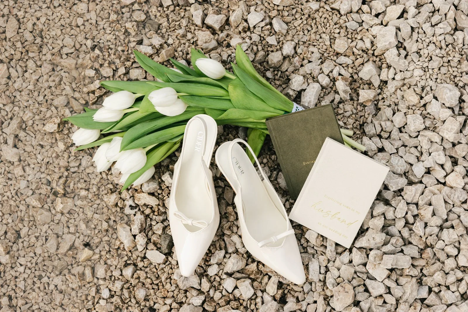 A pair of white high-heeled shoes, a bouquet of white tulips with green leaves, and two books with gold lettering on the covers are arranged on a bed of small rocks.