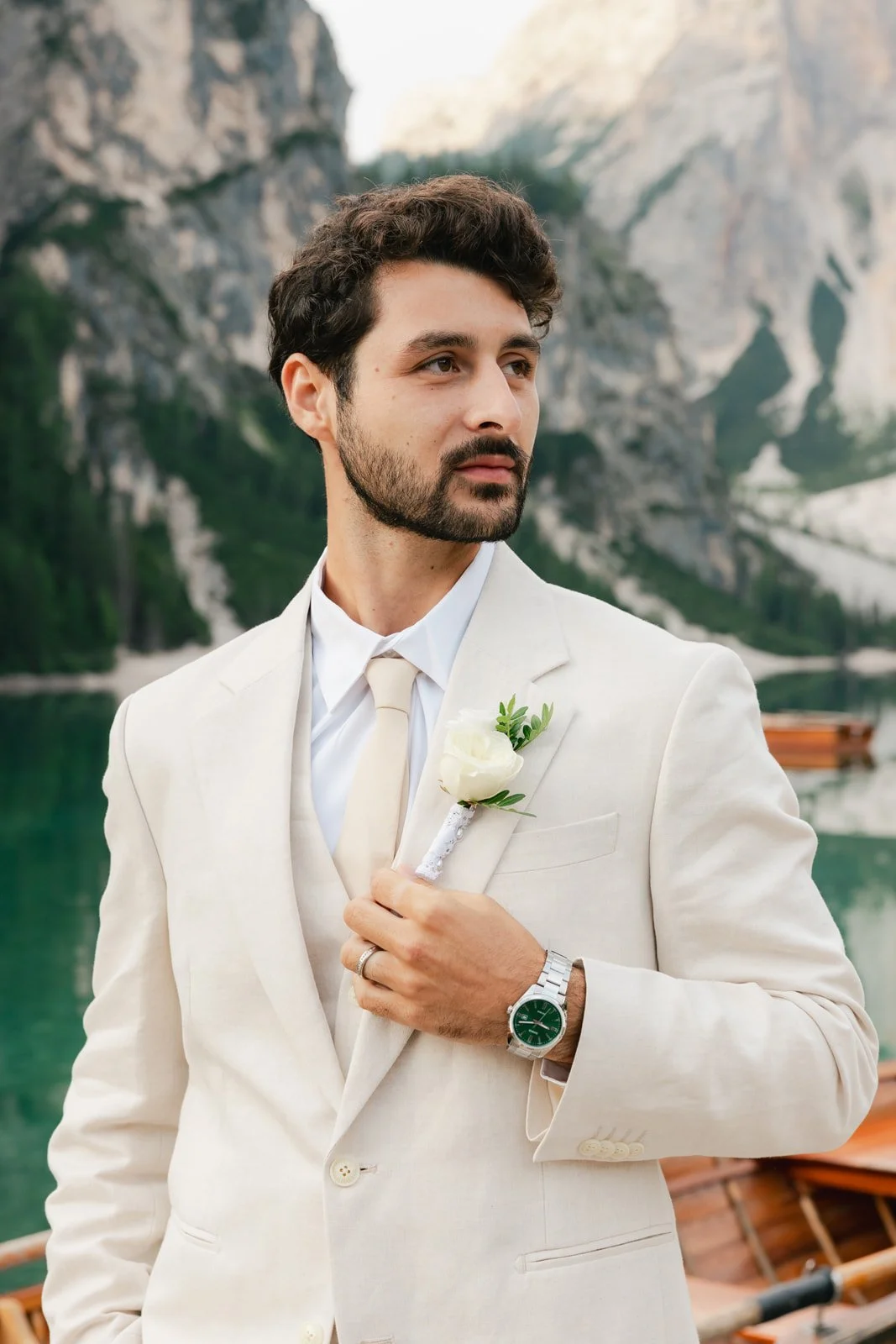 Man dressed in a cream suit, gold tie, white shirt, with a boutonniere on his lapel, standing near a body of water with mountains in the background. Lago Di Braies in the Dolomites of Italy