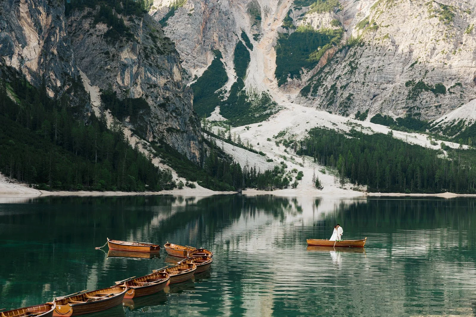 A mountain lake with several small wooden boats floating on calm water, surrounded by steep, rocky mountains and dense green forest in the background, with a couple standing in a boat on the right side. Lago Di Braies in the Dolomites of Italy