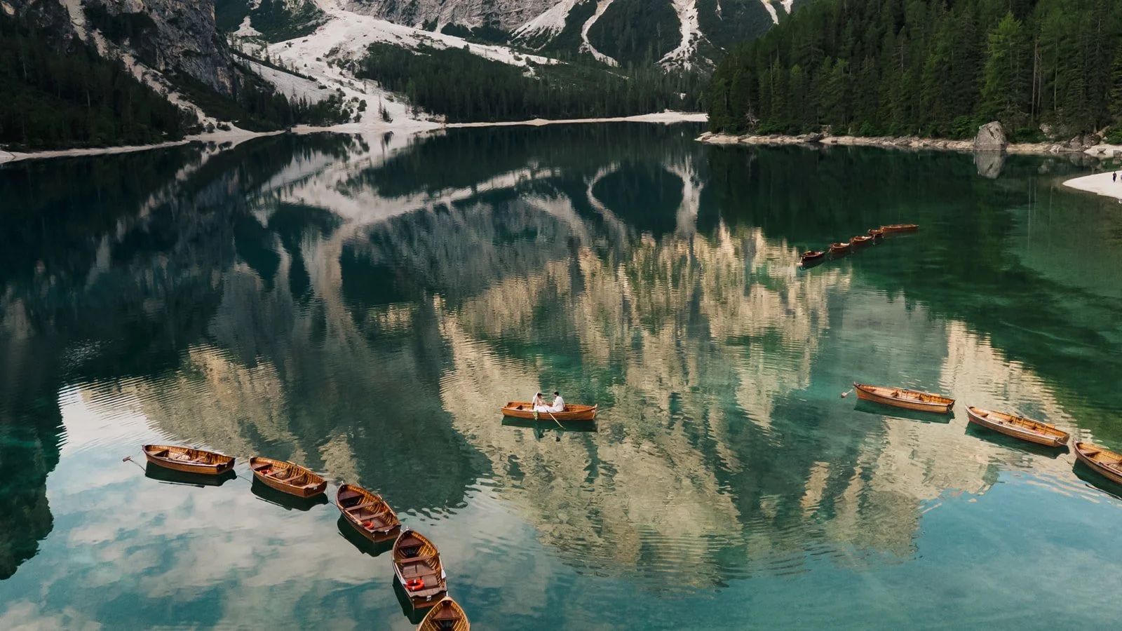 Scenic photo of a calm lake surrounded by snow-capped mountains and dense evergreen forests, with wooden boats floating on the water and two people sitting in one of the boats. Lago Di Braies in the Dolomites of Italy