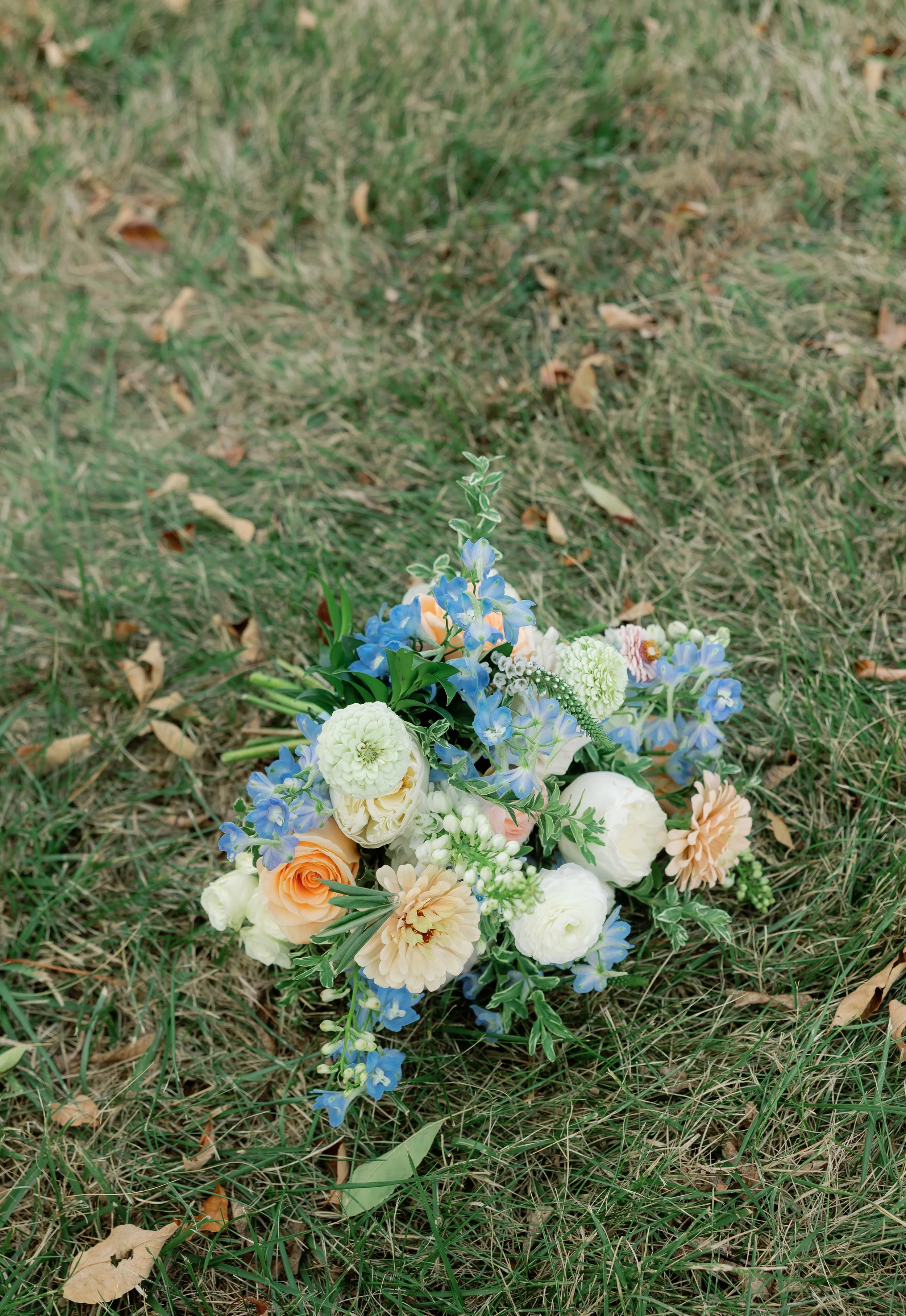 Callie’s pastel blue bridal bouquet rests gently on the ground, showcasing its white blooms, soft greenery, and peach accents. A beautifully styled floral moment perfect for brides searching for romantic, garden-inspired wedding bouquets.