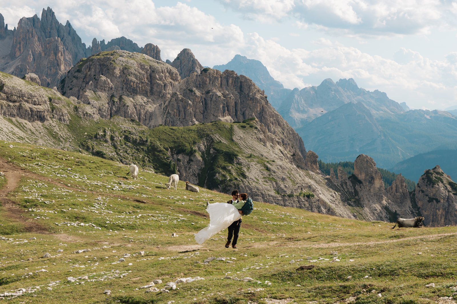 A couple in wedding attire embraces in a mountain meadow with grazing cattle and rocky peaks in the background.