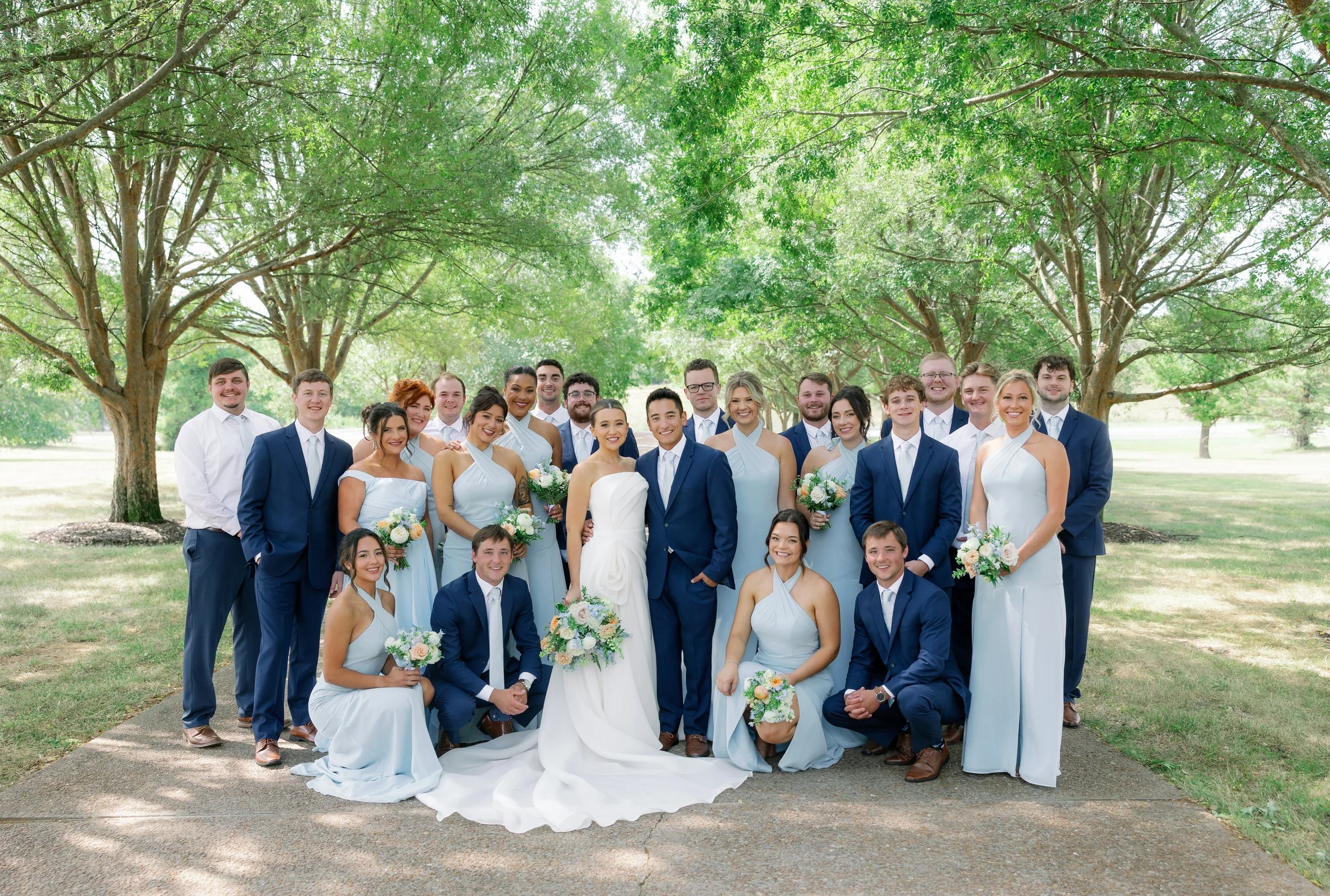 The entire bridal party gathered outdoors at The Callaway Jewel in Fulton, Missouri, dressed in soft pastel blue with coordinating white and peach florals. A bright summer backdrop highlighted the romantic color palette, beautifully captured by Jenni