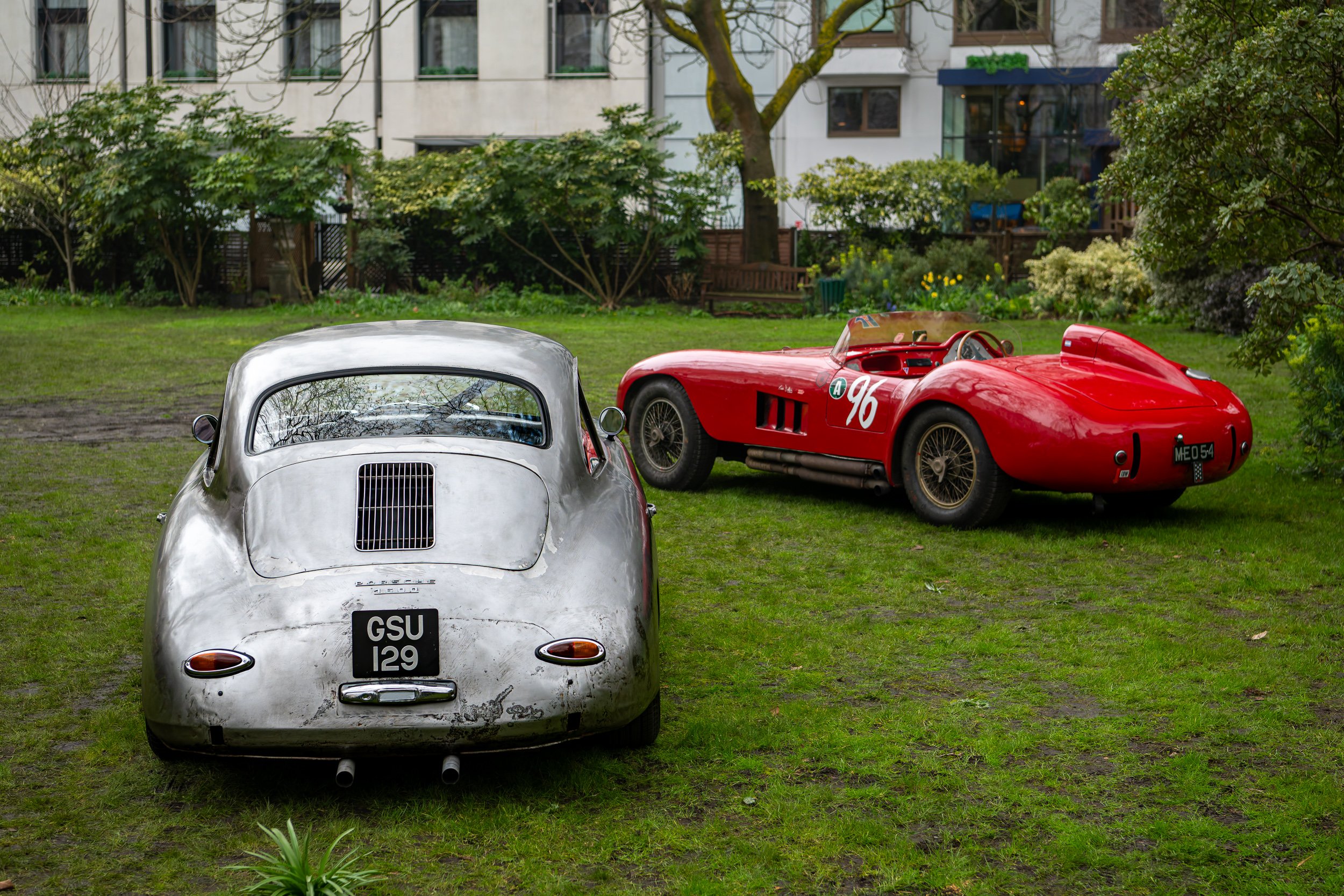 Two vintage sports cars, one silver with a rounded shape and the other red with an open cockpit, parked on a grassy area with trees and a building in the background.