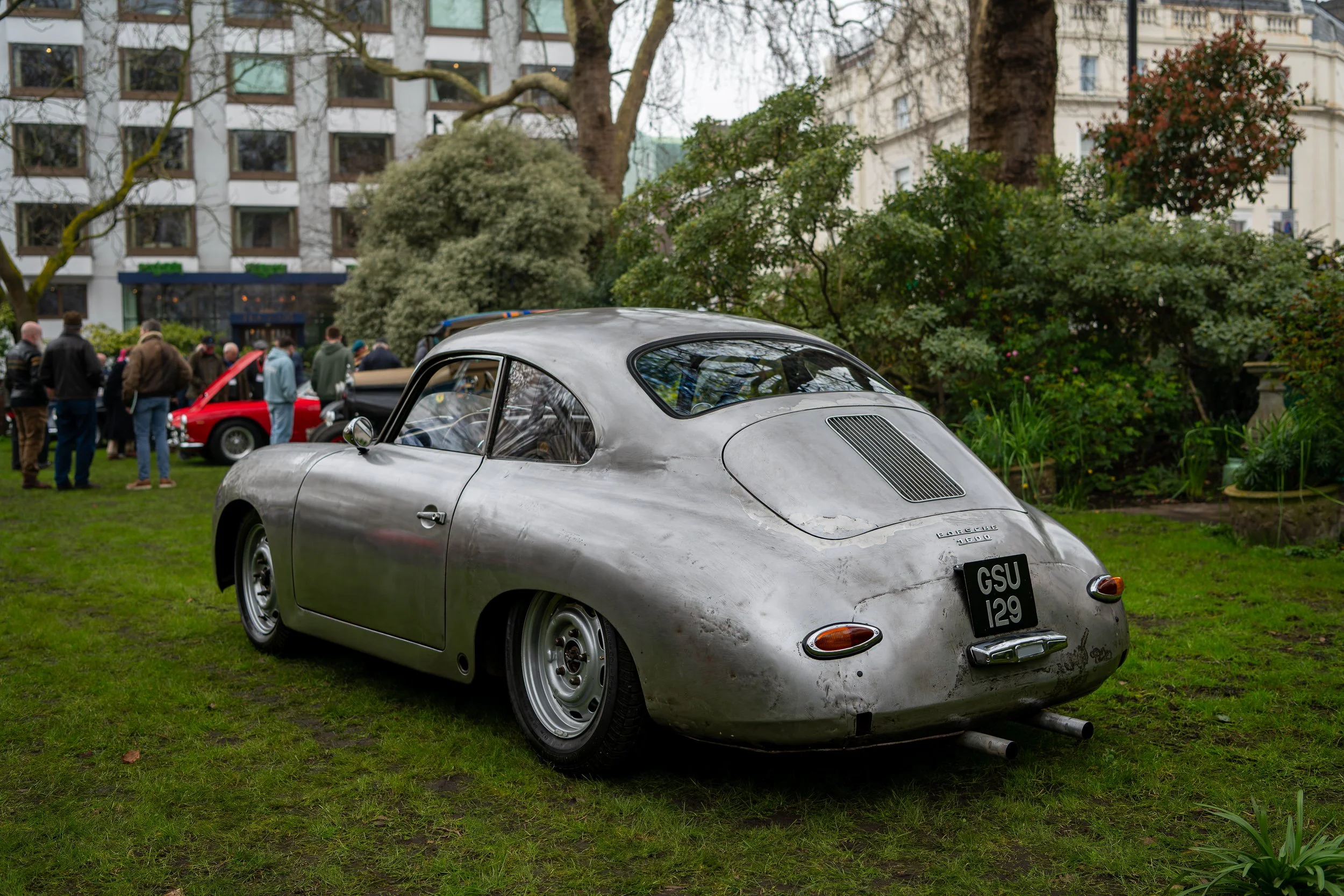 A vintage silver car, likely a Porsche 356, parked on a grassy area at a car show with people and other classic cars in the background.