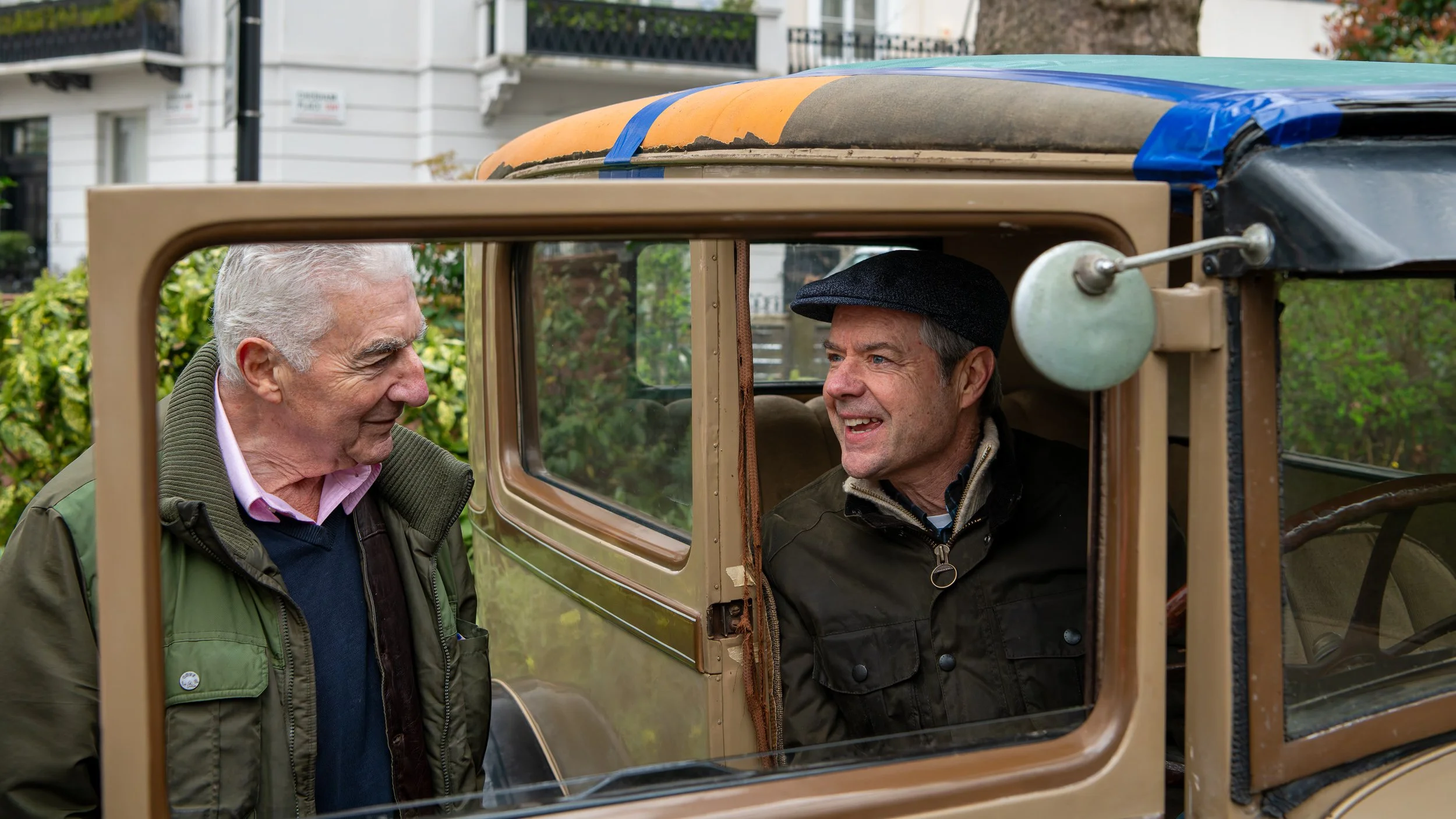 Two older men smiling and talking inside a vintage car with the window rolled down.