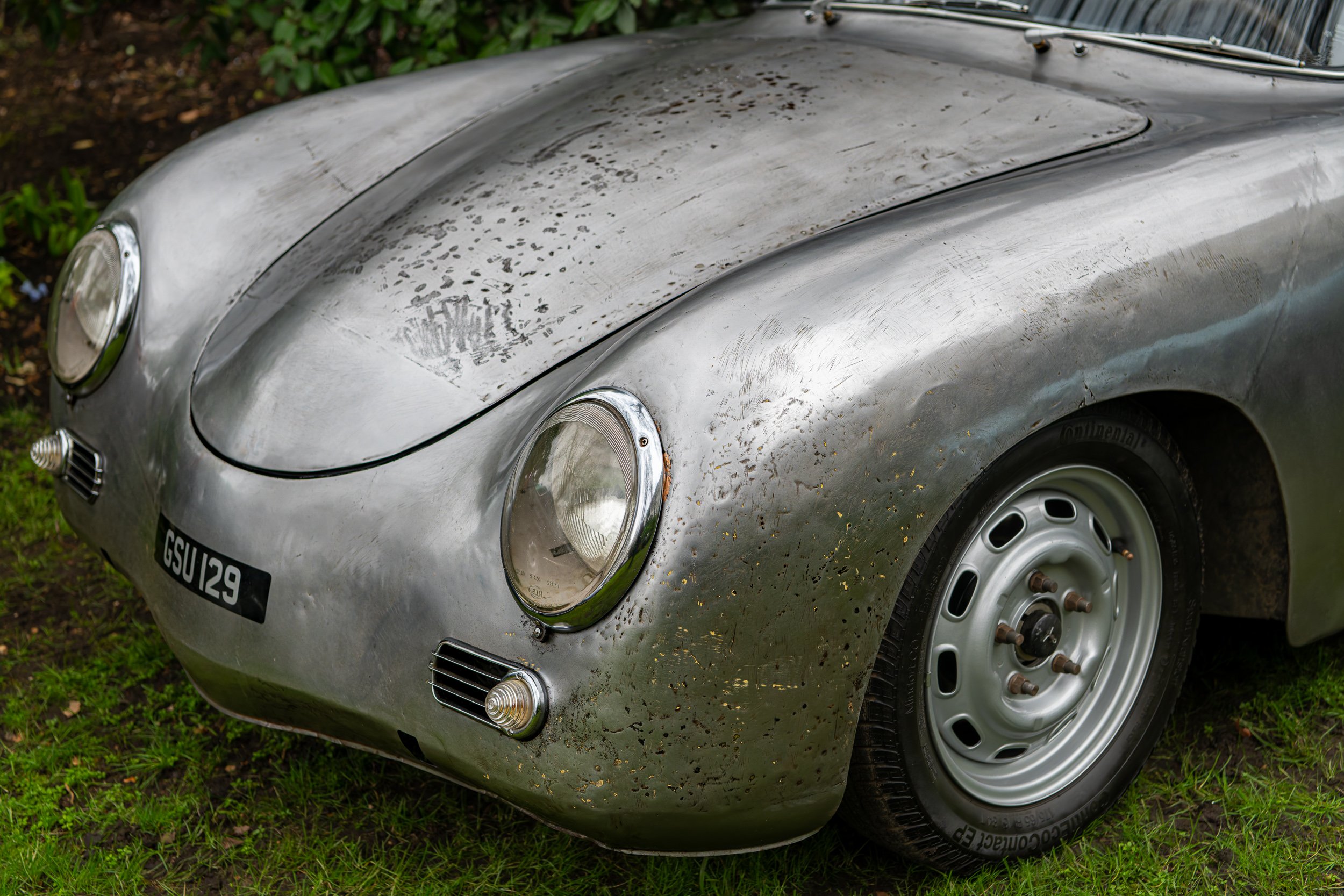 Front view of a vintage silver car with rusty spots, round headlights, and a black license plate reading GS1129, parked on grass.