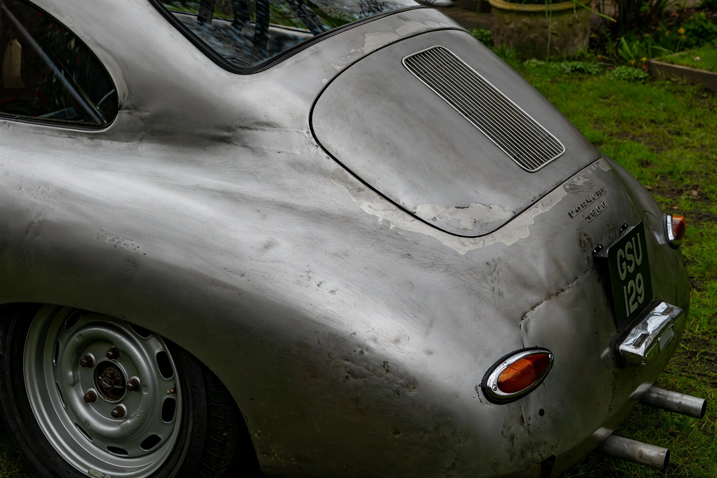 Rear view of a vintage silver sports car with visible dents and rust spots, parked on grass. The car has a black license plate with white lettering, twin exhaust pipes, and a vented engine hood.