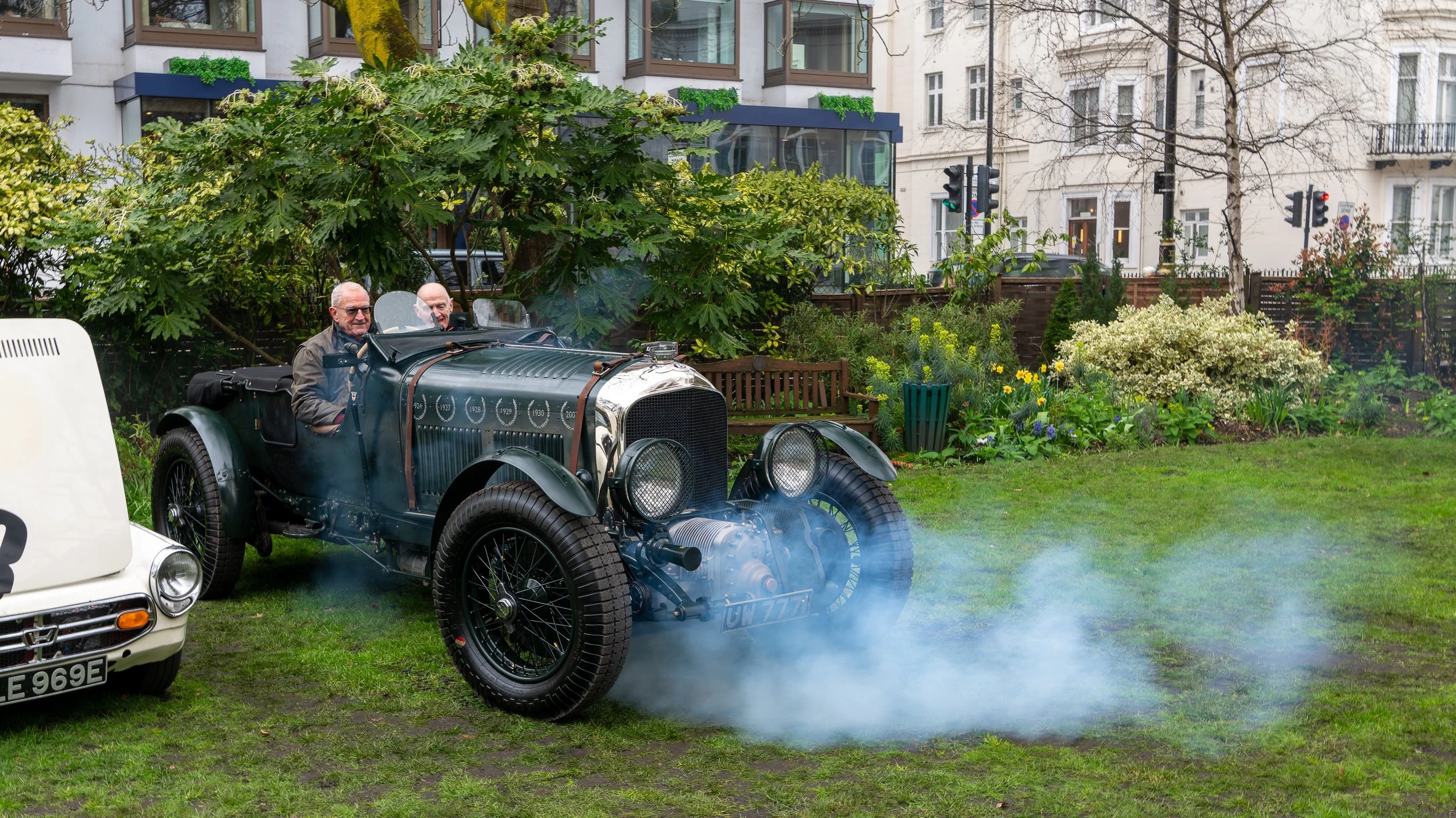Two elderly men are driving an antique race car on a lawn, with smoke coming from the back, in a garden with trees, bushes, and flowers in front of white buildings.