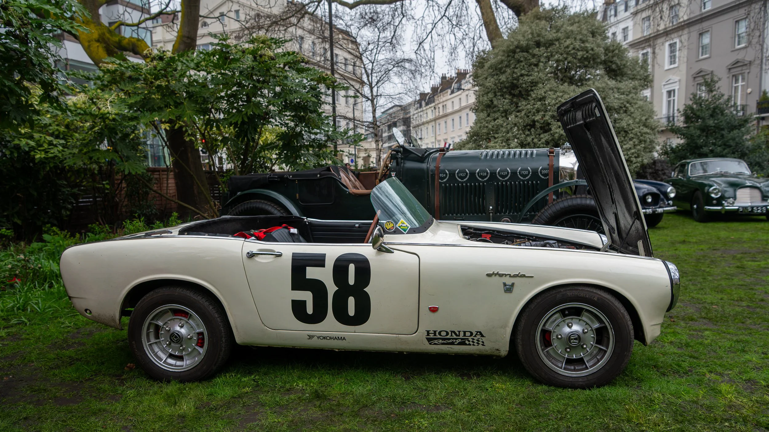A vintage white Honda racing car with the number 58 on the side, parked on grass with its hood open. Other classic cars are visible in the background among trees and buildings.