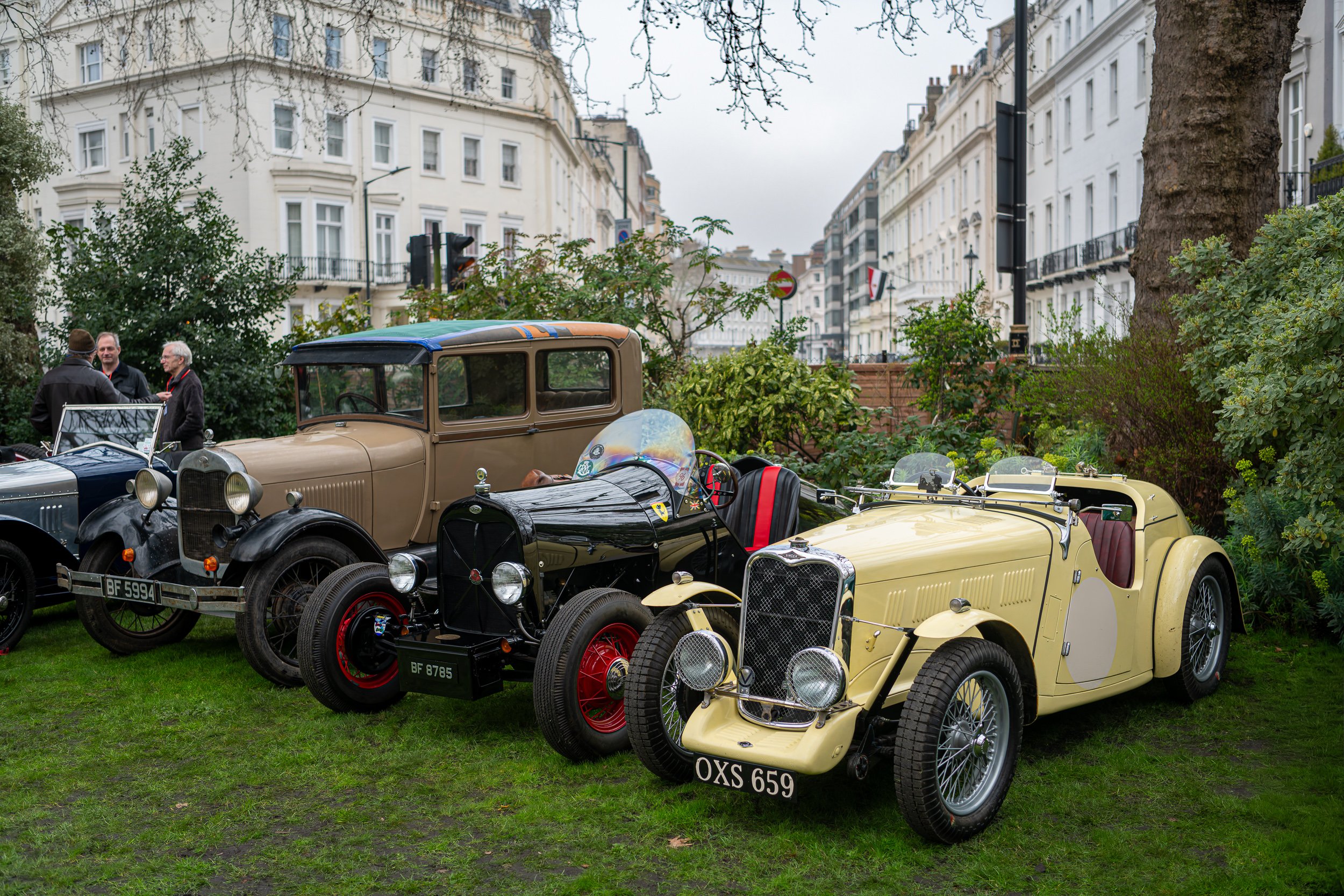Several vintage cars parked on a grassy area with three people talking in the background, surrounded by trees and white buildings.