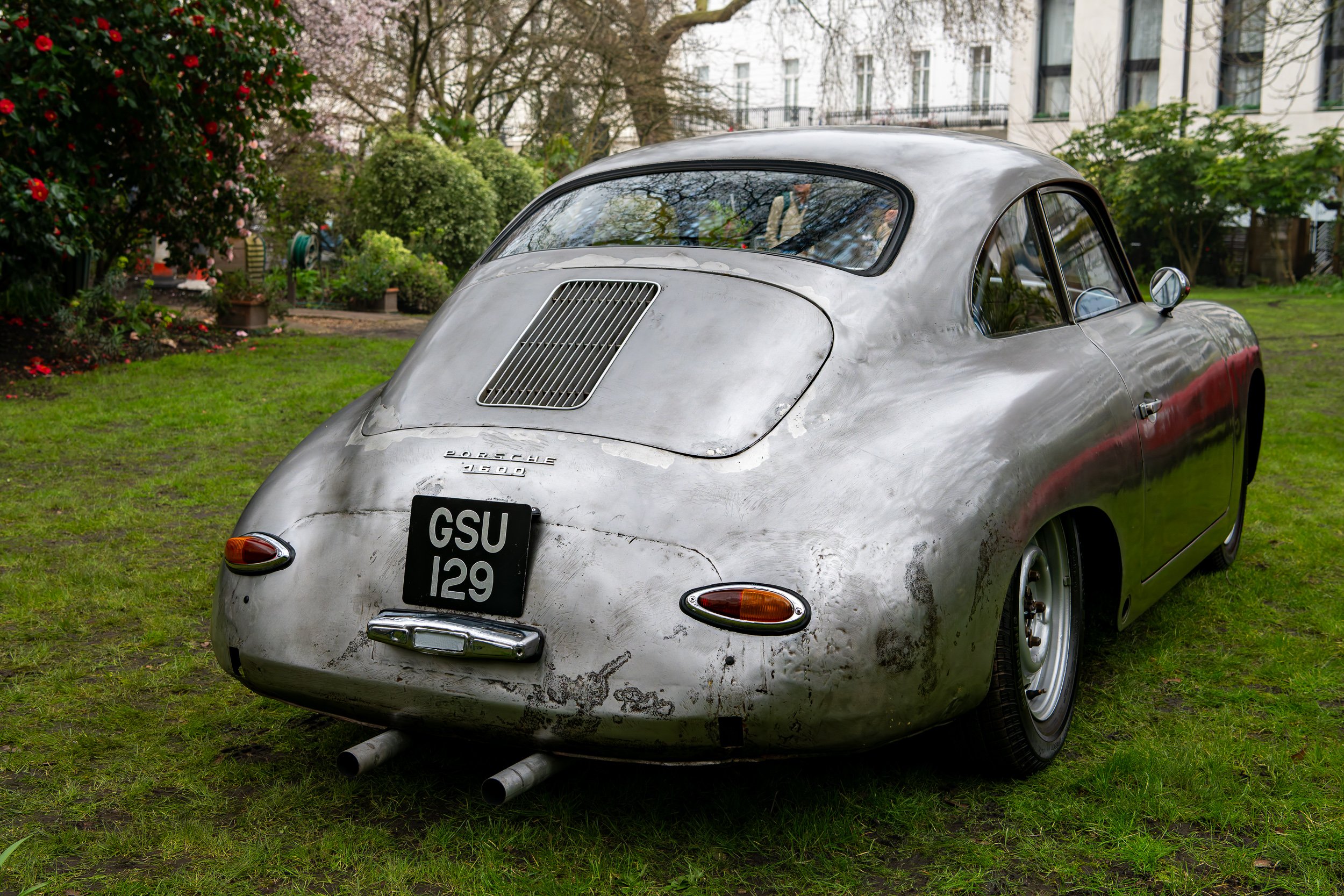 A vintage silver Porsche 356 parked on a grassy area with trees and buildings in the background.