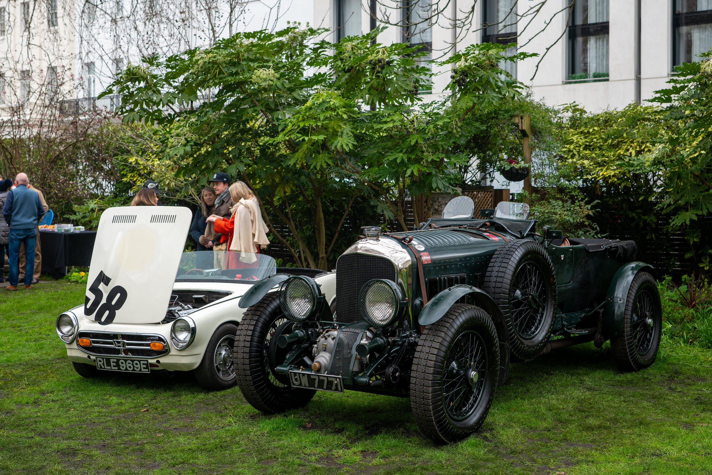 Two vintage cars, a white race car with a large number 58 on its hood and a black classic car, parked on a grassy area at a car show with several people gathered around. There are trees and a white building in the background.