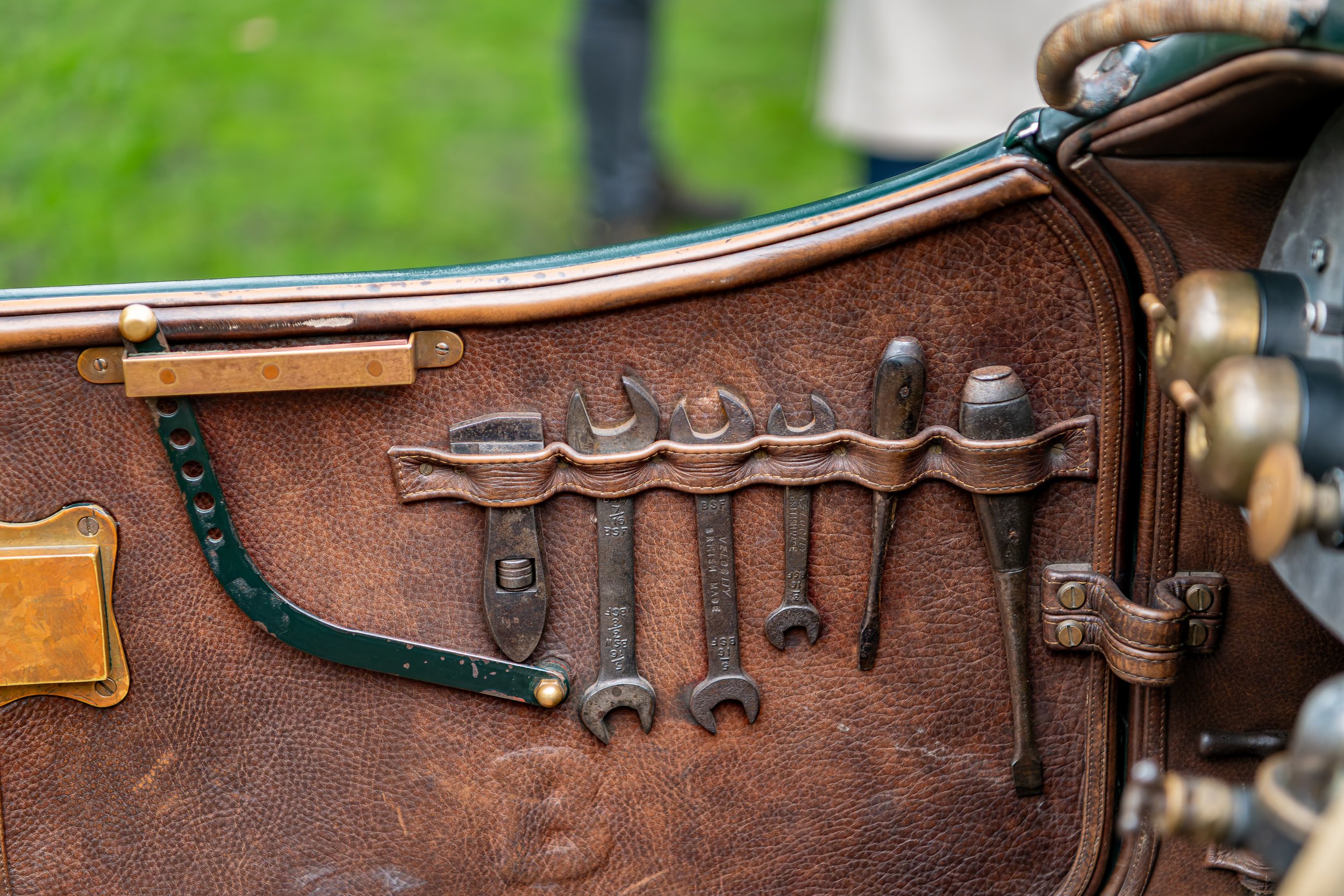 An old leather tool roll on a vintage motorcycle's fuel tank, holding various hand tools including wrenches, screwdrivers, and a set of pliers, with a grassy outdoor background.