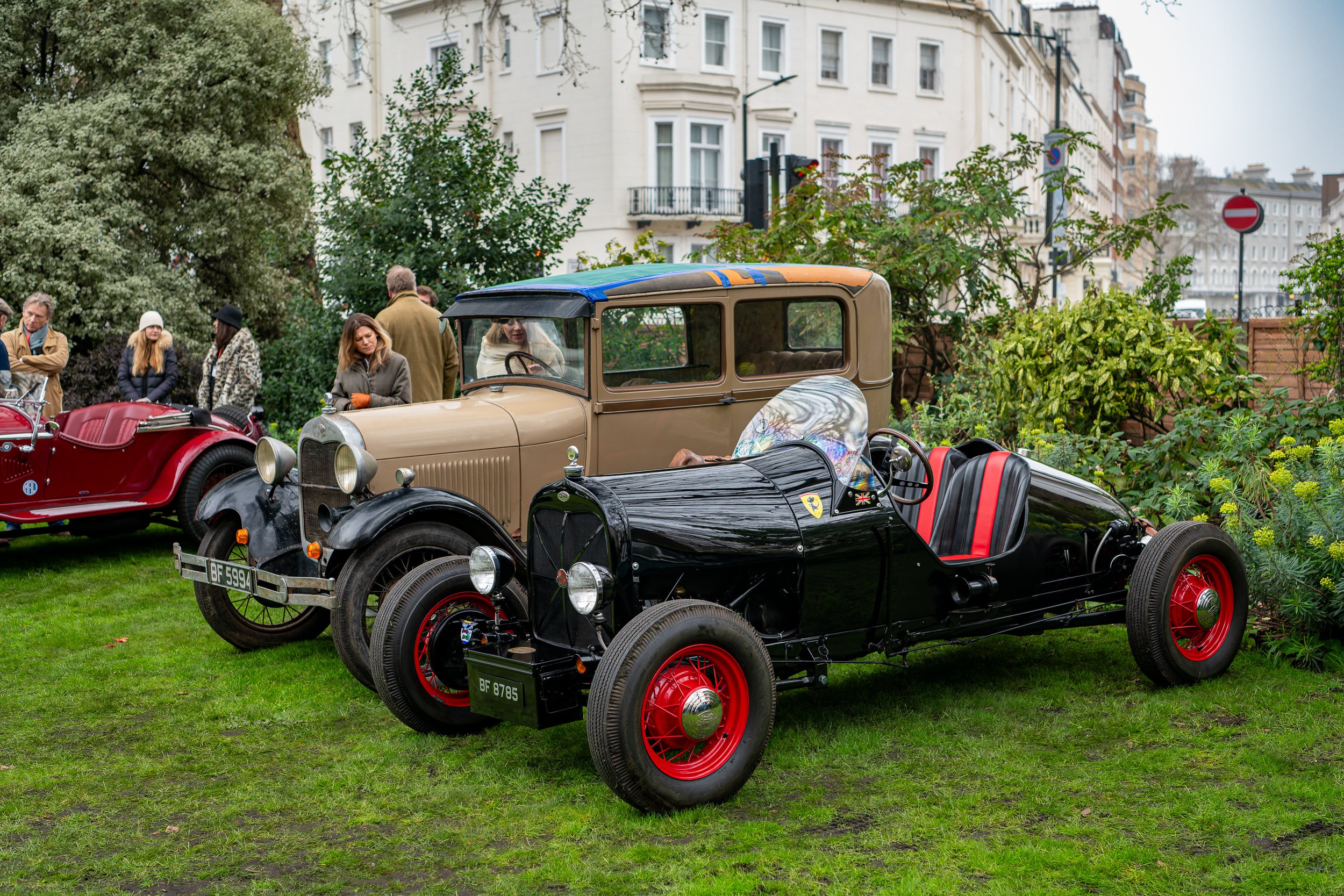 Three vintage cars parked on a grassy area during a car show, with a group of people looking at them and city buildings in the background.