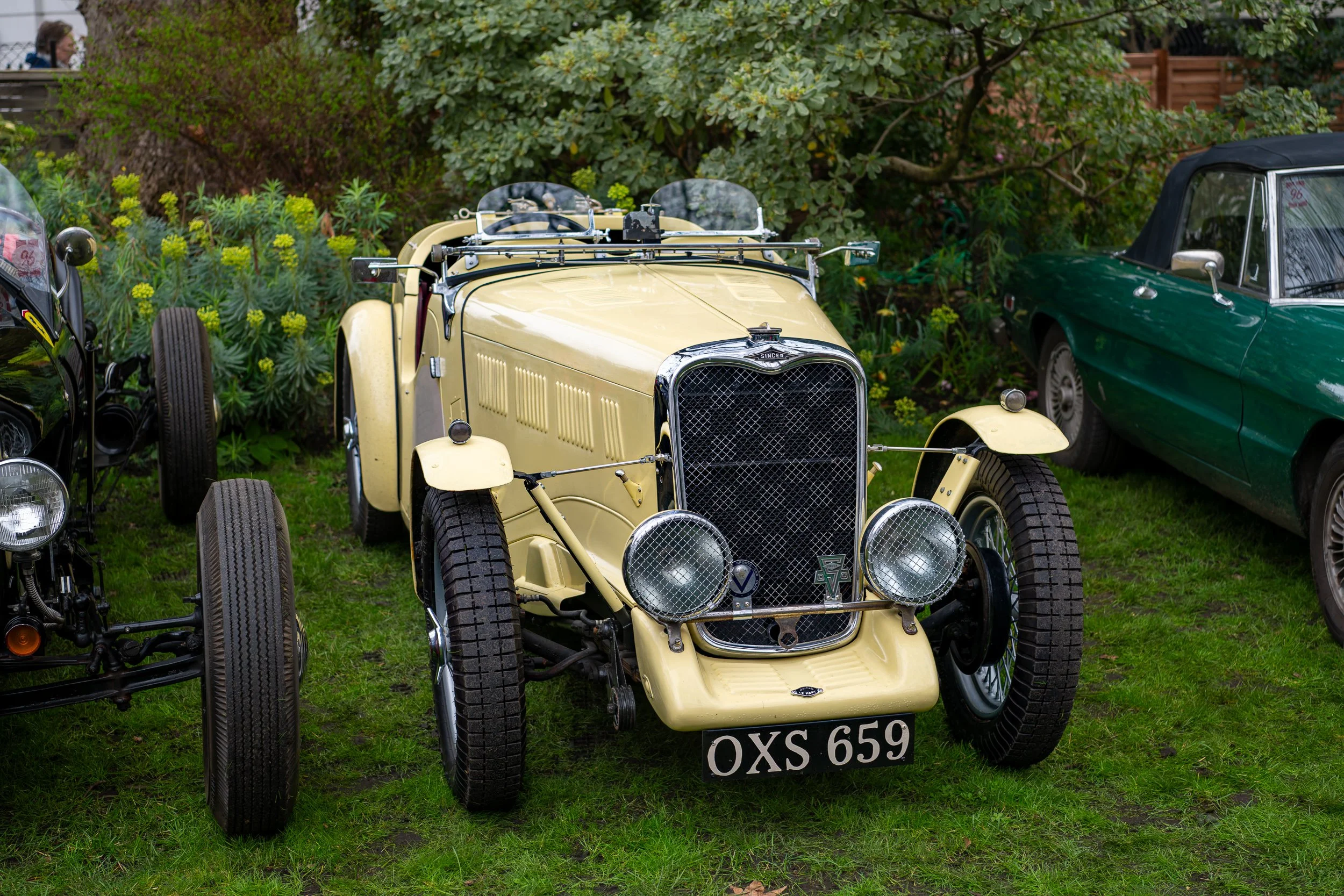 A vintage yellow open-top car with a front grille, round headlights, and a black license plate reading 'OXS 659,' parked on grass in a garden surrounded by other classic cars and green plants.