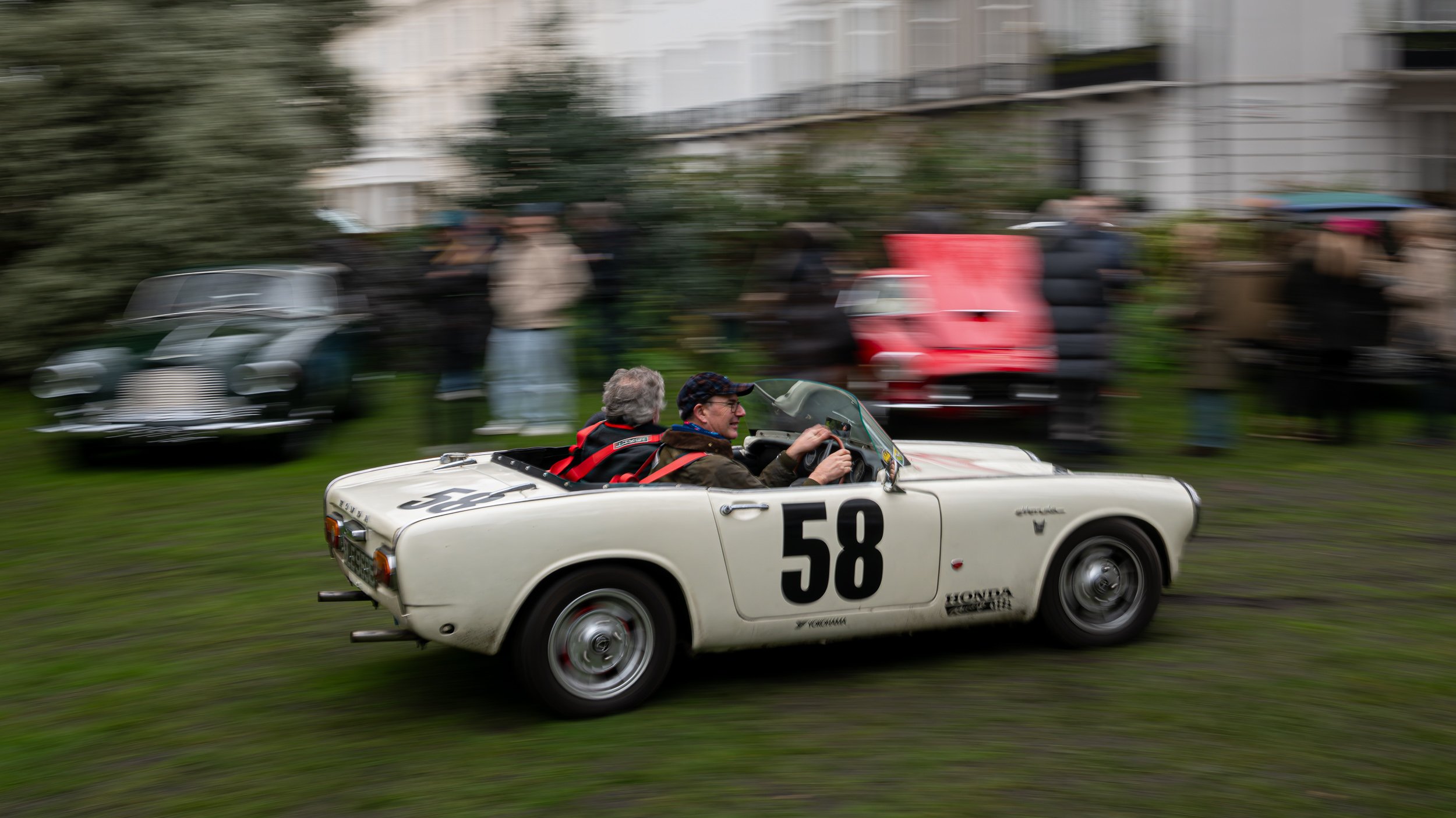A white vintage racing car with the number 58 on its side, driven by a man and a passenger in a crowd of spectators during a race or event.