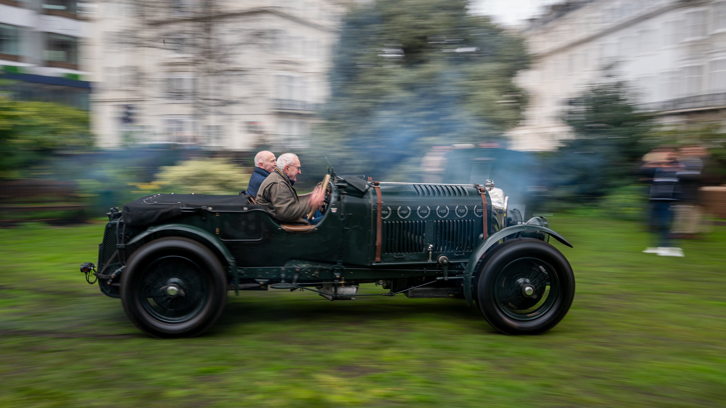 An old vintage black car with two men inside, moving fast with a blurred background of buildings and people, on a grassy area.