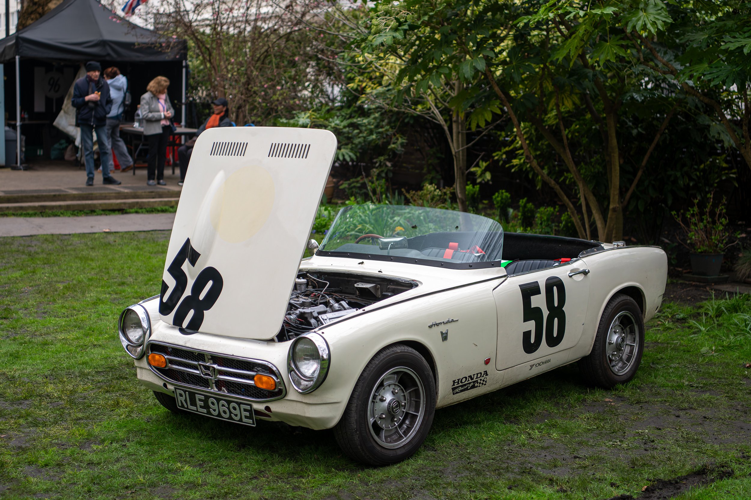 A vintage white Honda sports car with open hood and racing number 58 on the side, parked on a grassy area during an outdoor event with people in the background.