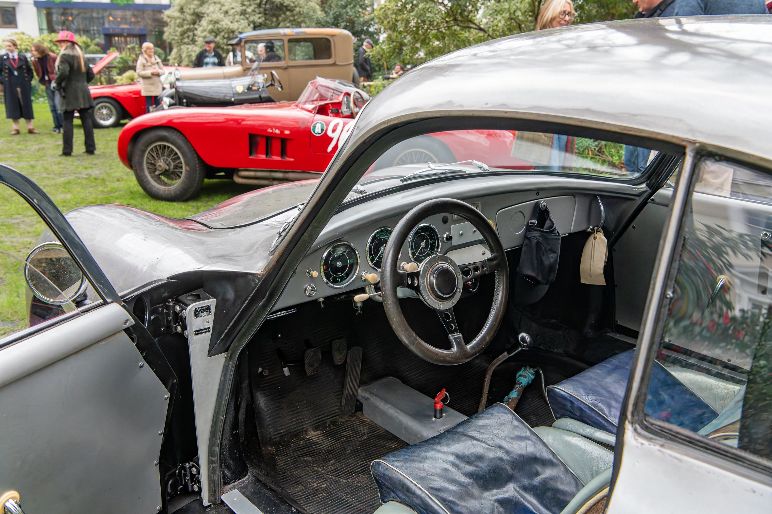 Interior of a vintage race car with a steering wheel, speedometer gauges, and racing gear, at a classic car show with people and other vintage cars visible outside.
