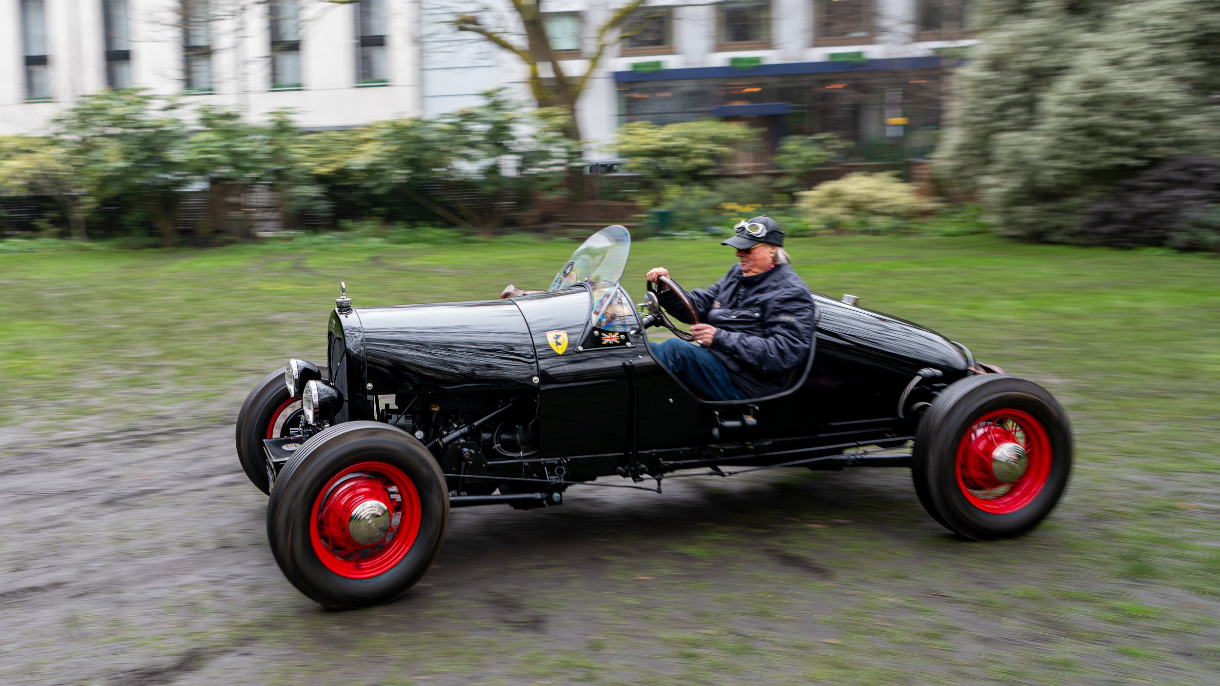 A man driving a vintage black race car with red wheels on a grassy area, with a blurred building and trees in the background.