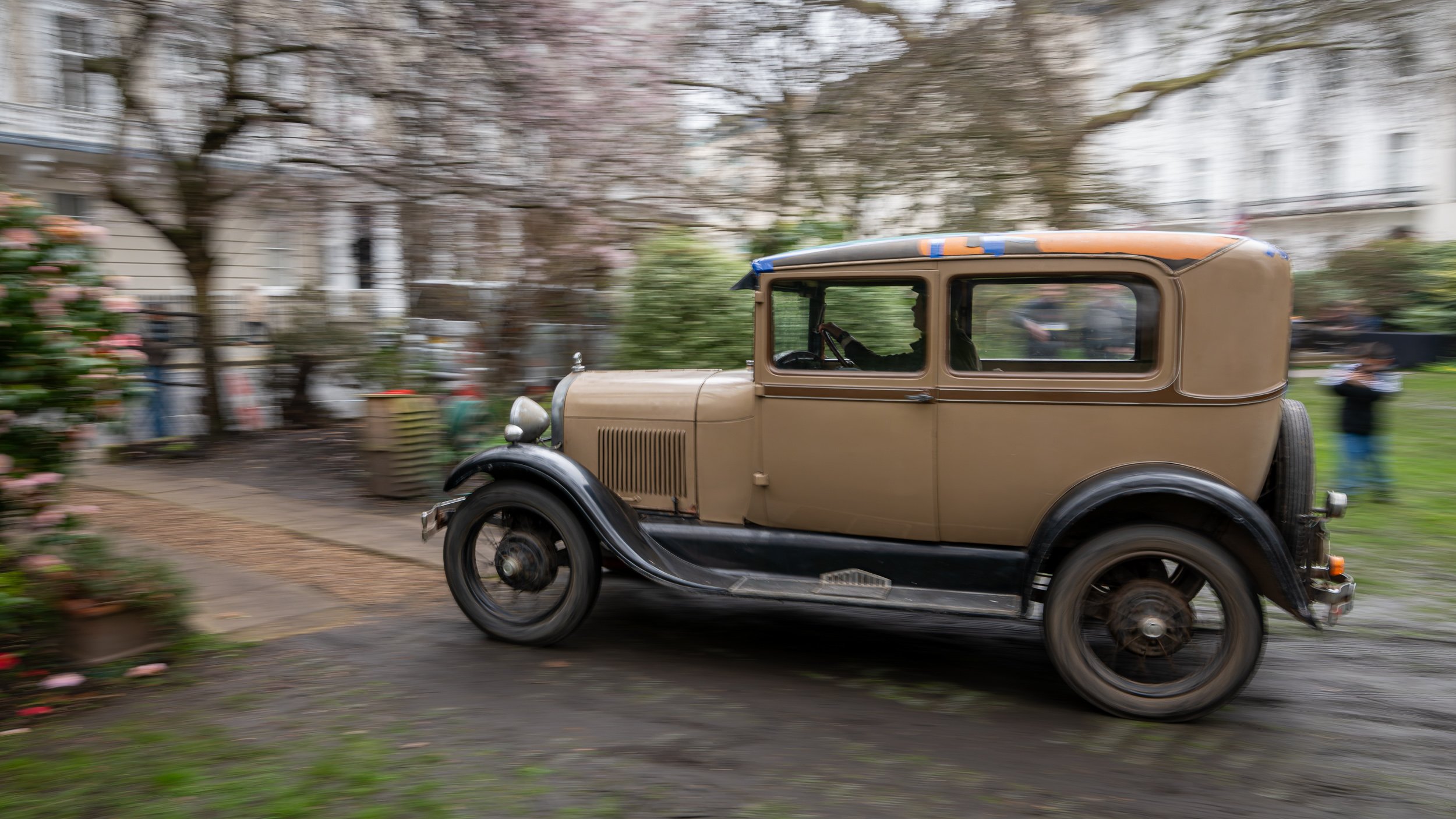 A vintage brown and black car driving through a park with trees and flowers, motion blurred background.