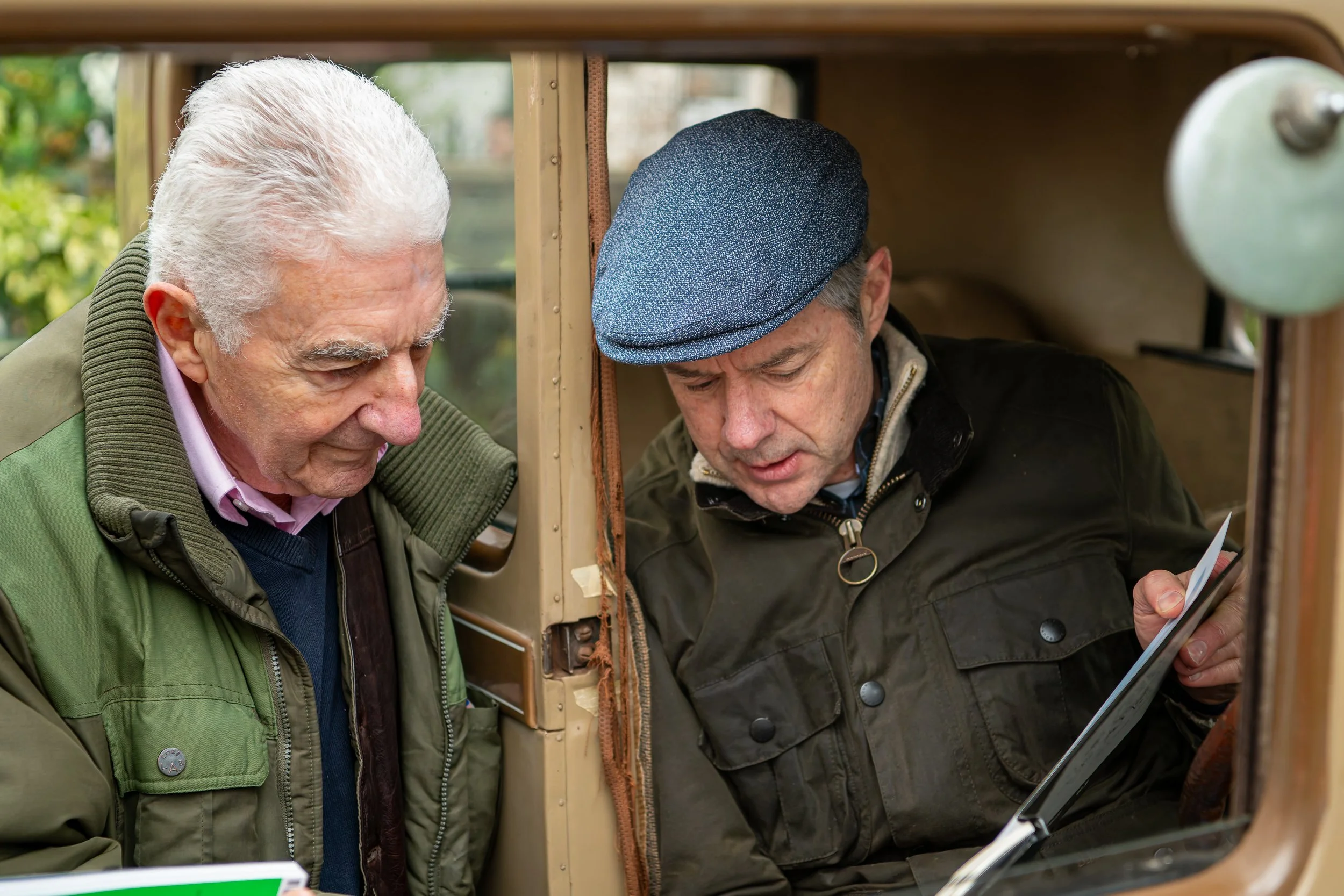 Two older men looking at a tablet inside a vintage car, one with a gray cap and black jacket, the other with white hair and a green jacket.