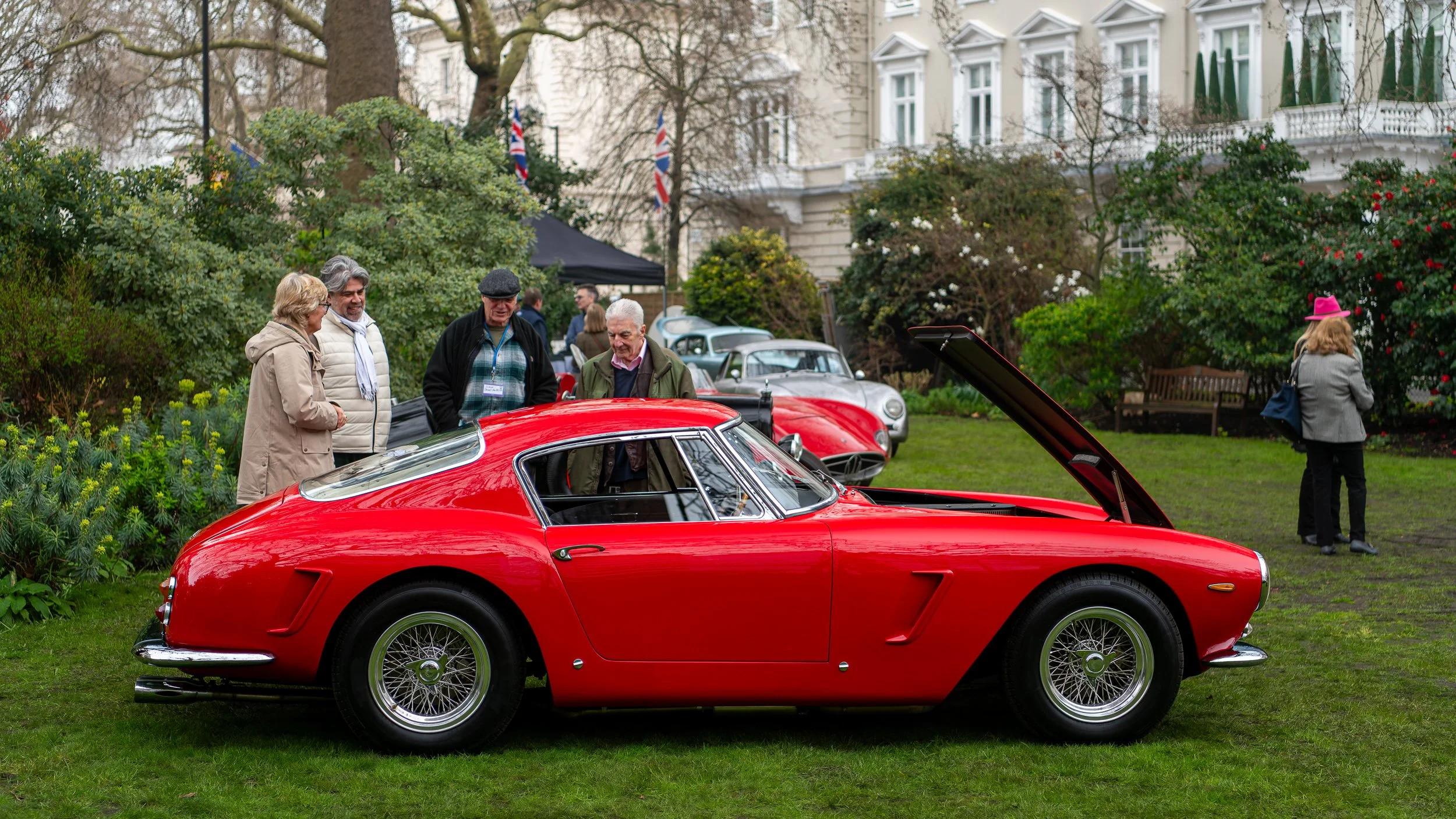 A red vintage sports car on display at an outdoor classic car show with four people examining it, and other classic cars and people in the background in a garden setting.