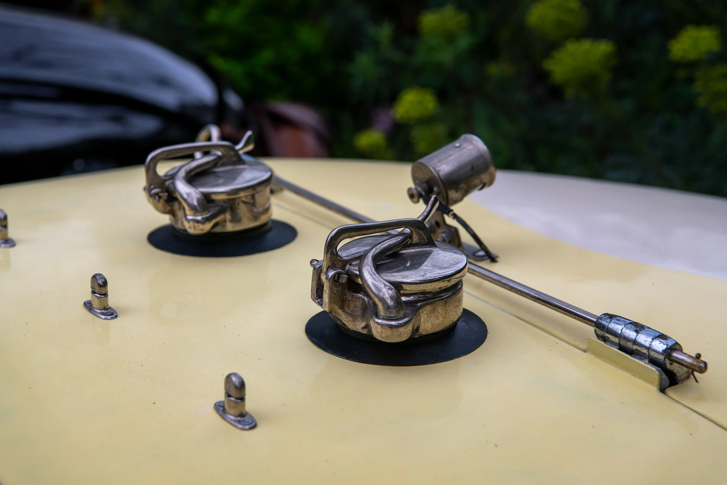 Close-up of two metallic hoods with handles and a mechanical component on the hood of a vintage yellow car, with a blurred green background.