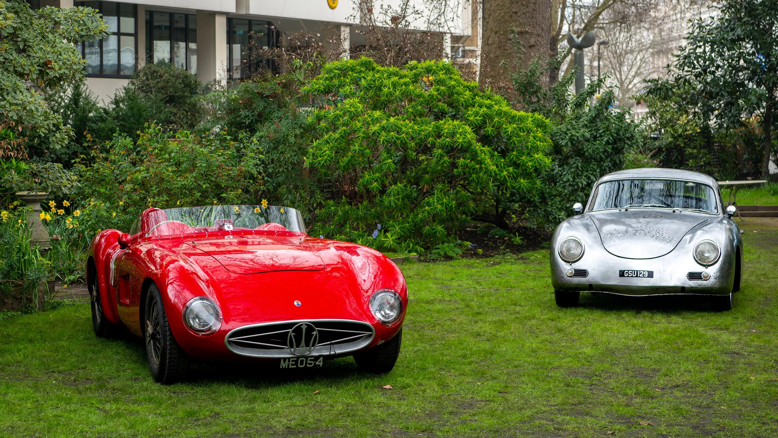 A red vintage race car and a silver classic Porsche parked on a grassy lawn with bushes and trees in the background.