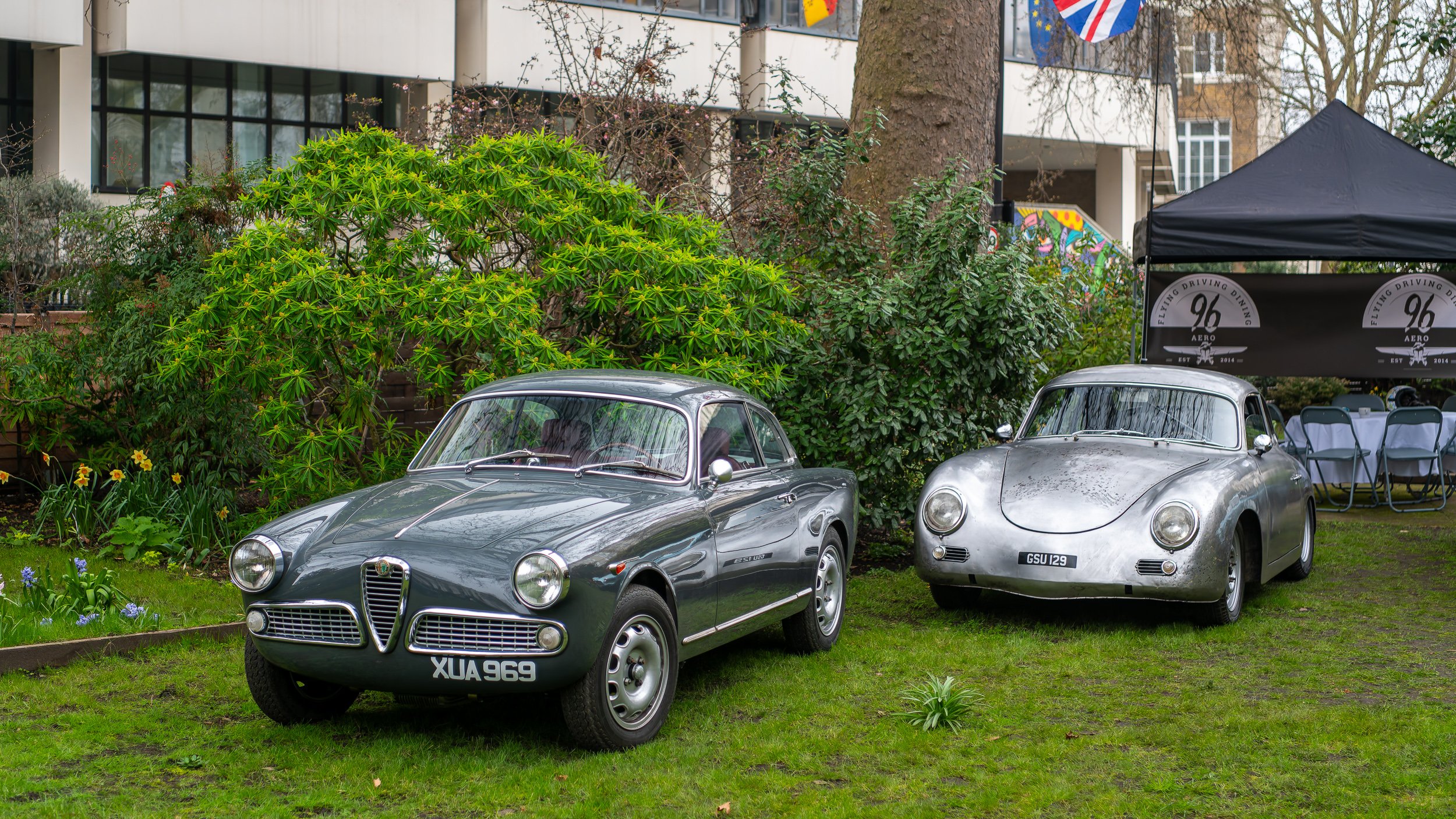 Two vintage cars, one dark gray and one silver, parked on a grassy lawn near bushes and trees, with a black canopy tent and outdoor seating in the background.