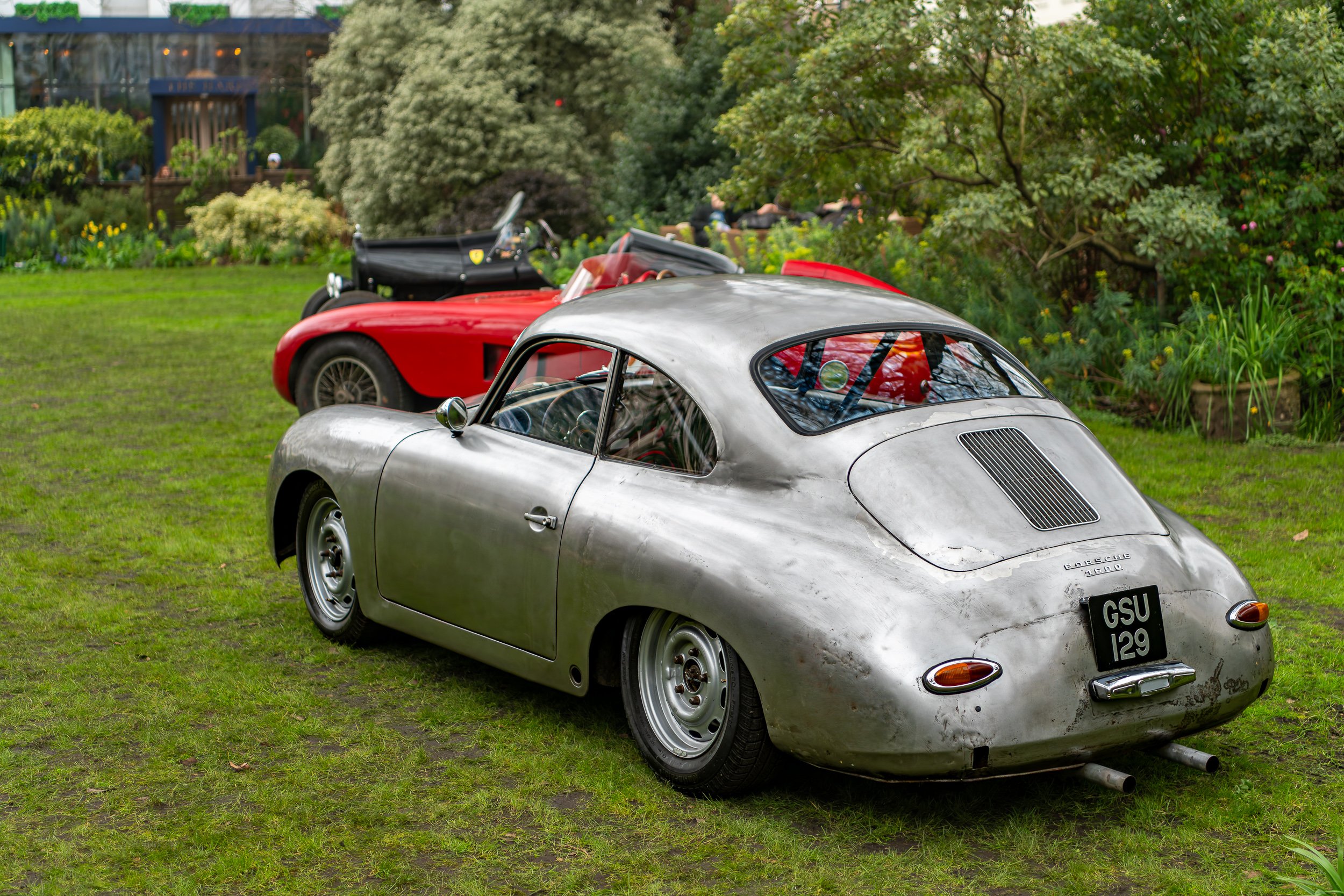 A vintage silver Porsche 356 with a weathered exterior is parked on grass, with a red convertible sports car and a black vintage car in the background, surrounded by lush greenery.