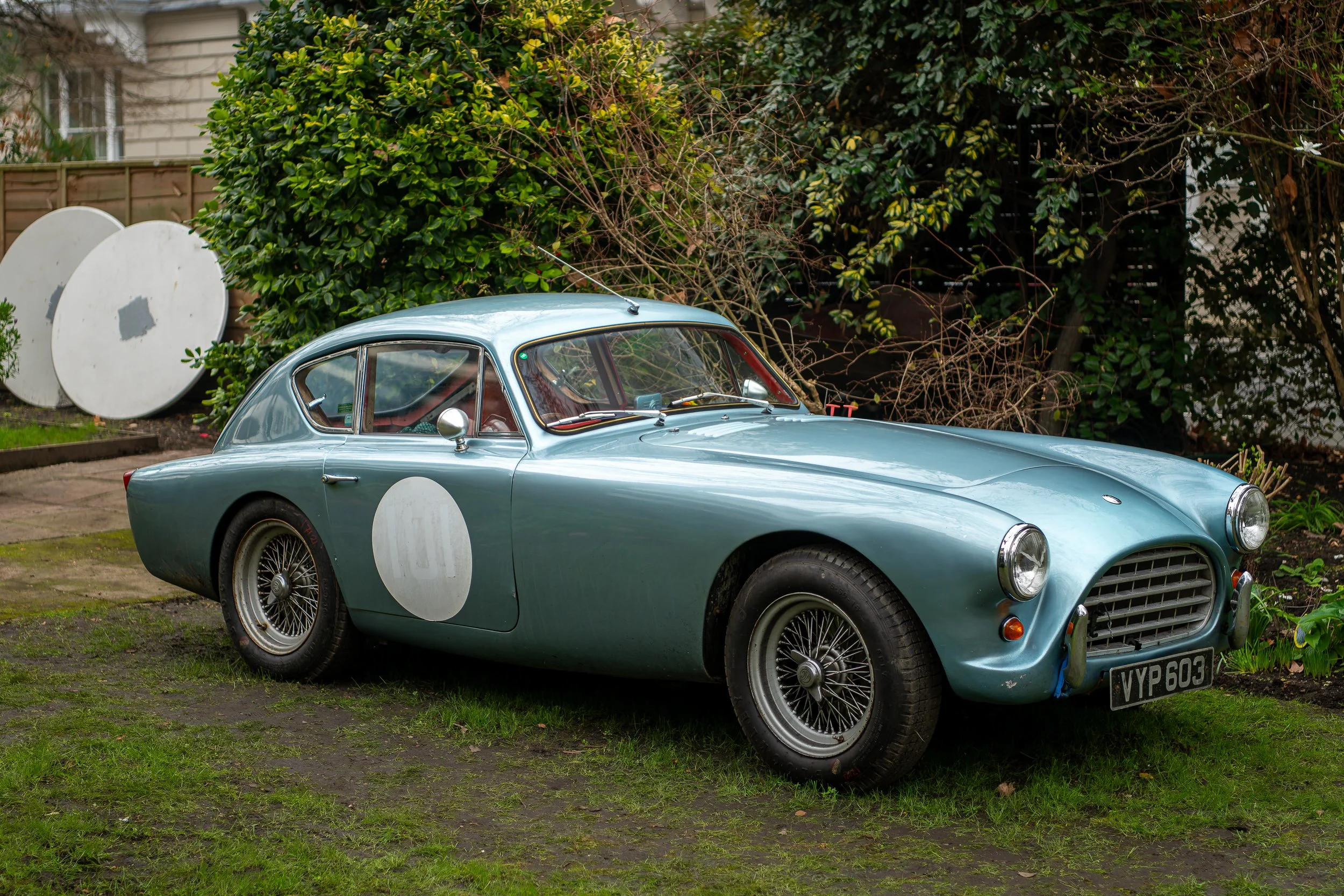 A vintage light blue race car with white circles on the door and hood, parked on grass in a garden. The car features wire-spoke wheels, round headlights, and a UK license plate reading 'VYP 603.'