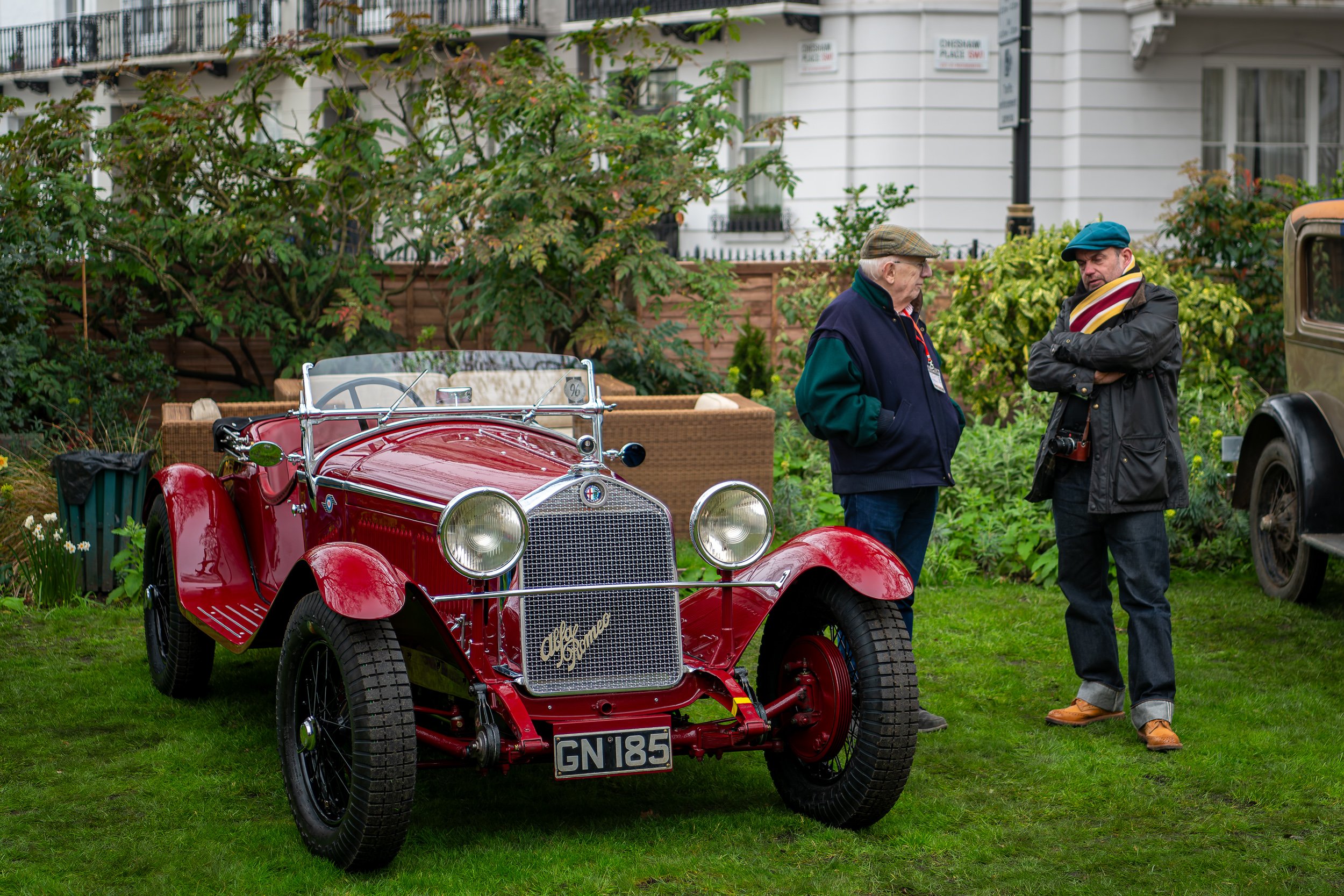 Two men standing on a lawn talking near a vintage red car, with a garden and a white house in the background.