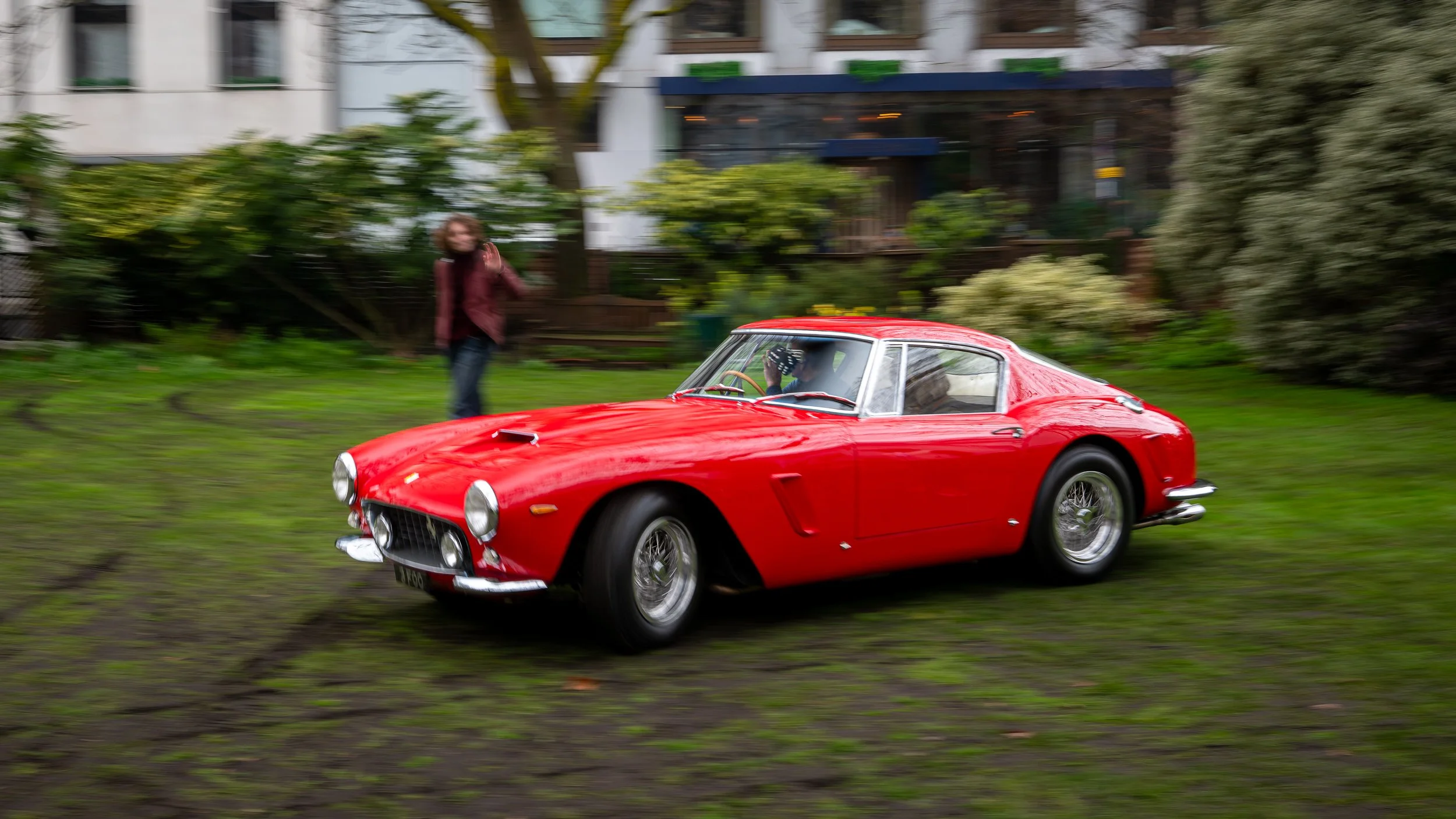 A red vintage car parked on a grassy area with a woman in the background waving.