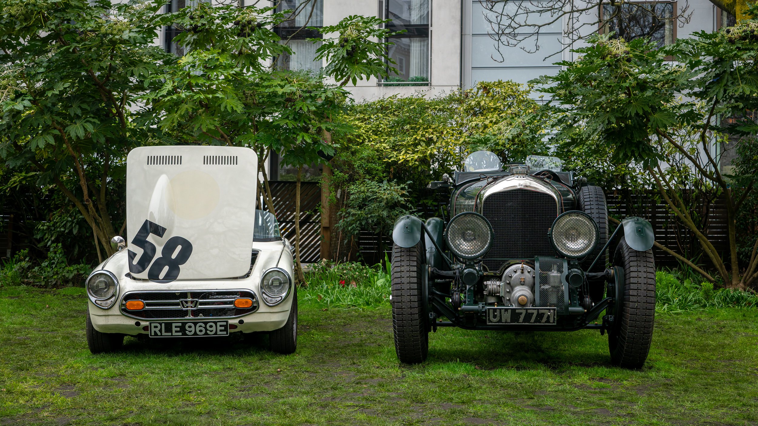 Two vintage cars, one with a white body and a racing number 58 on its hood cover, and one black with exposed engine parts, are parked side by side on grass in front of greenery and a building.